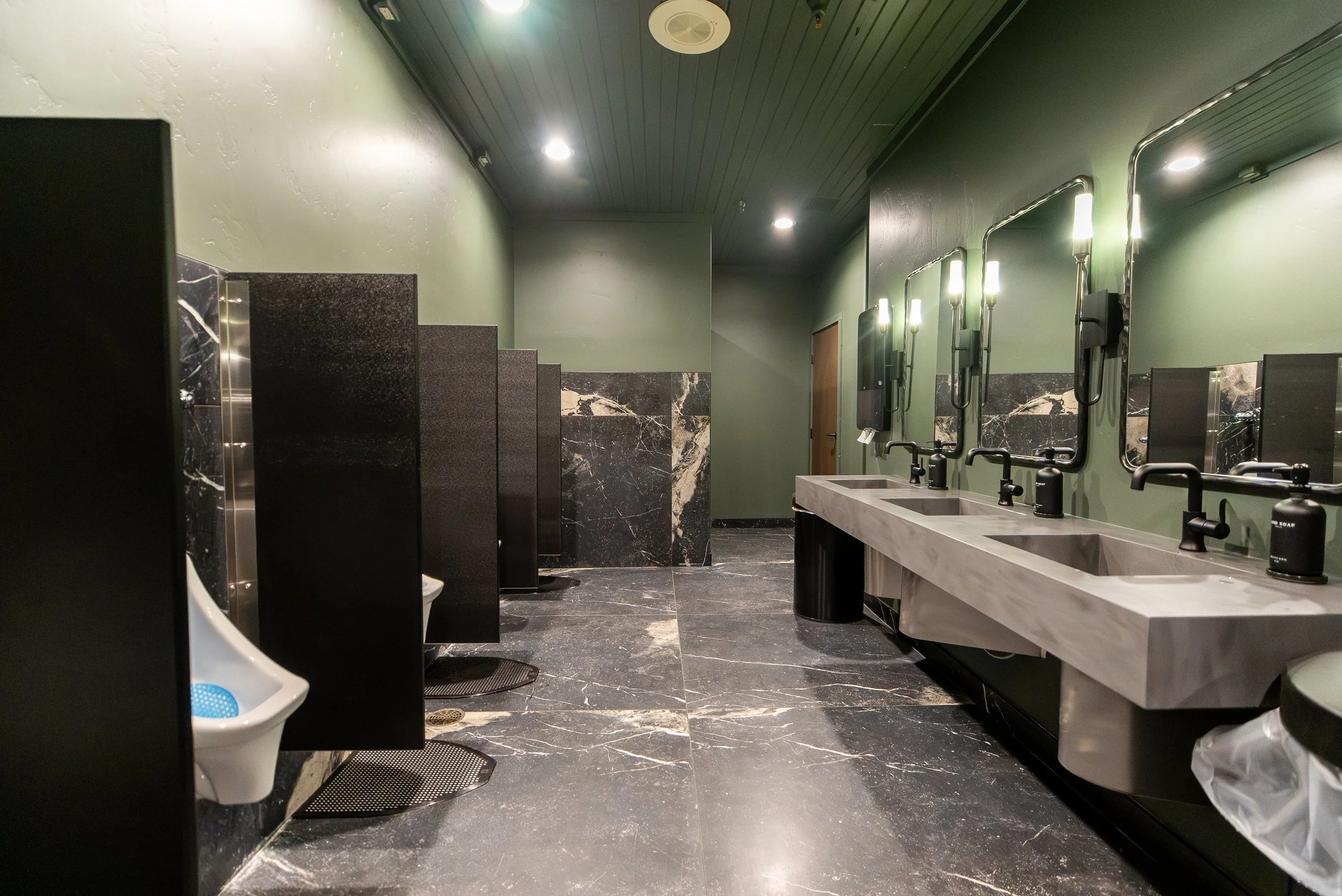 Public men's restroom with urinals on the left and sinks with mirrors on the right, tiled black floor, green walls, black marble accents, and ceiling lights.