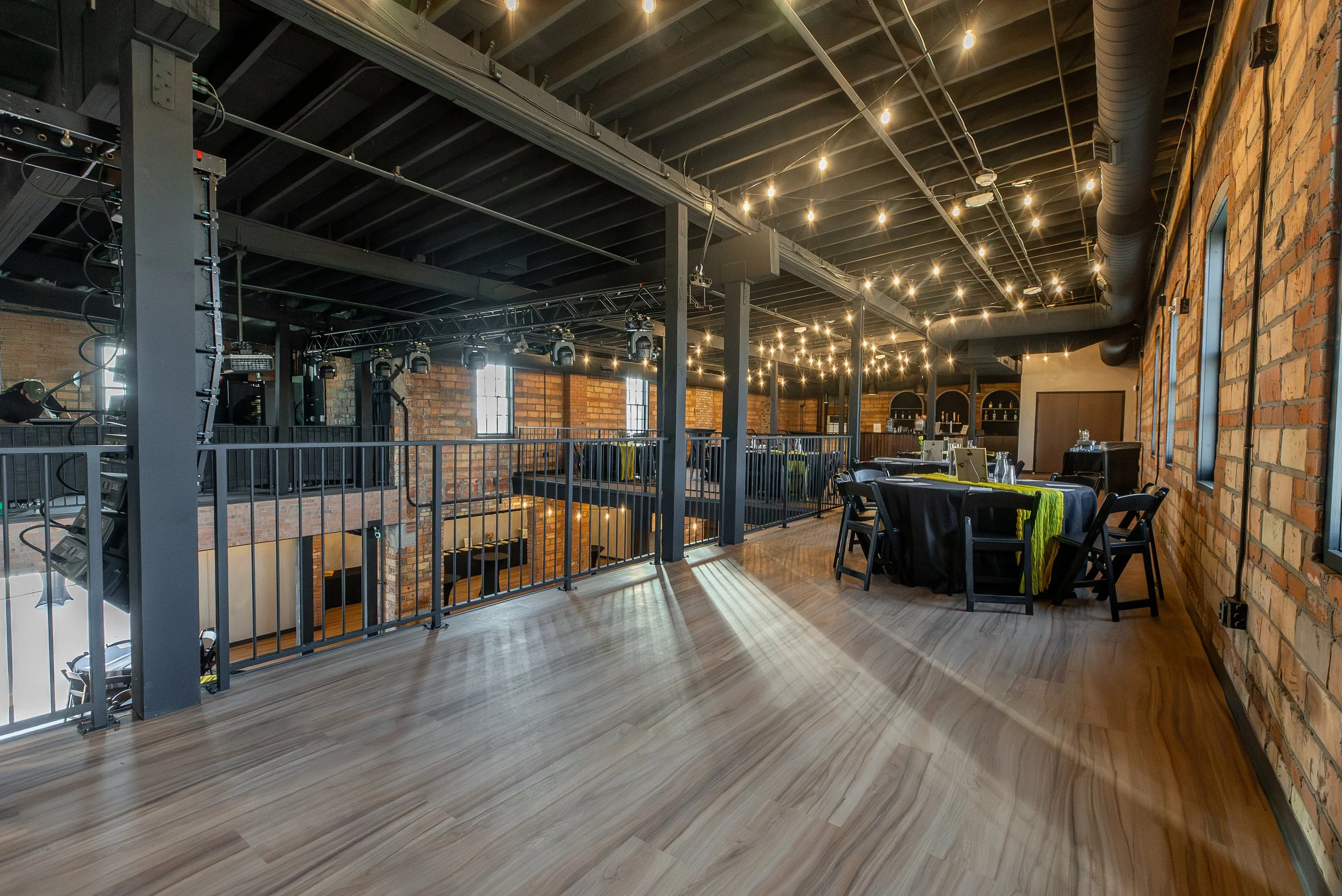 An interior of a modern industrial-style restaurant or event space with exposed brick walls, string lights on the ceiling, and tables with black chairs and yellow-green cloths.