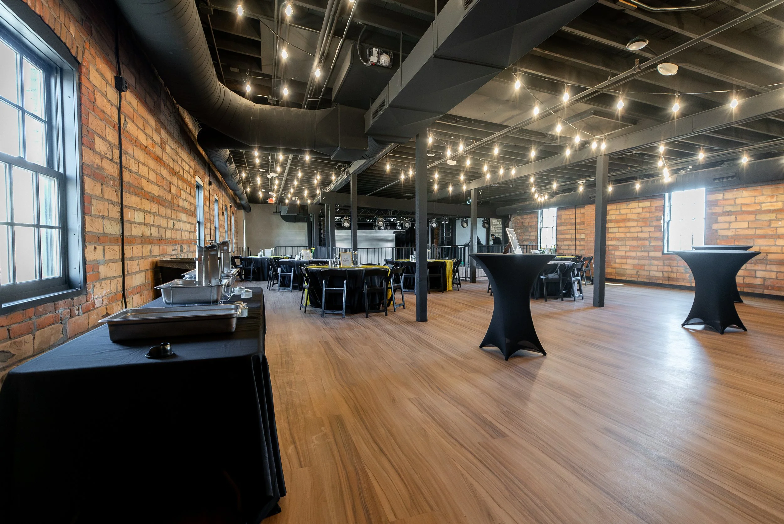 Interior of a modern event space or restaurant with exposed brick walls, large windows, wooden floors, string lights on the ceiling, and tables set for dining.