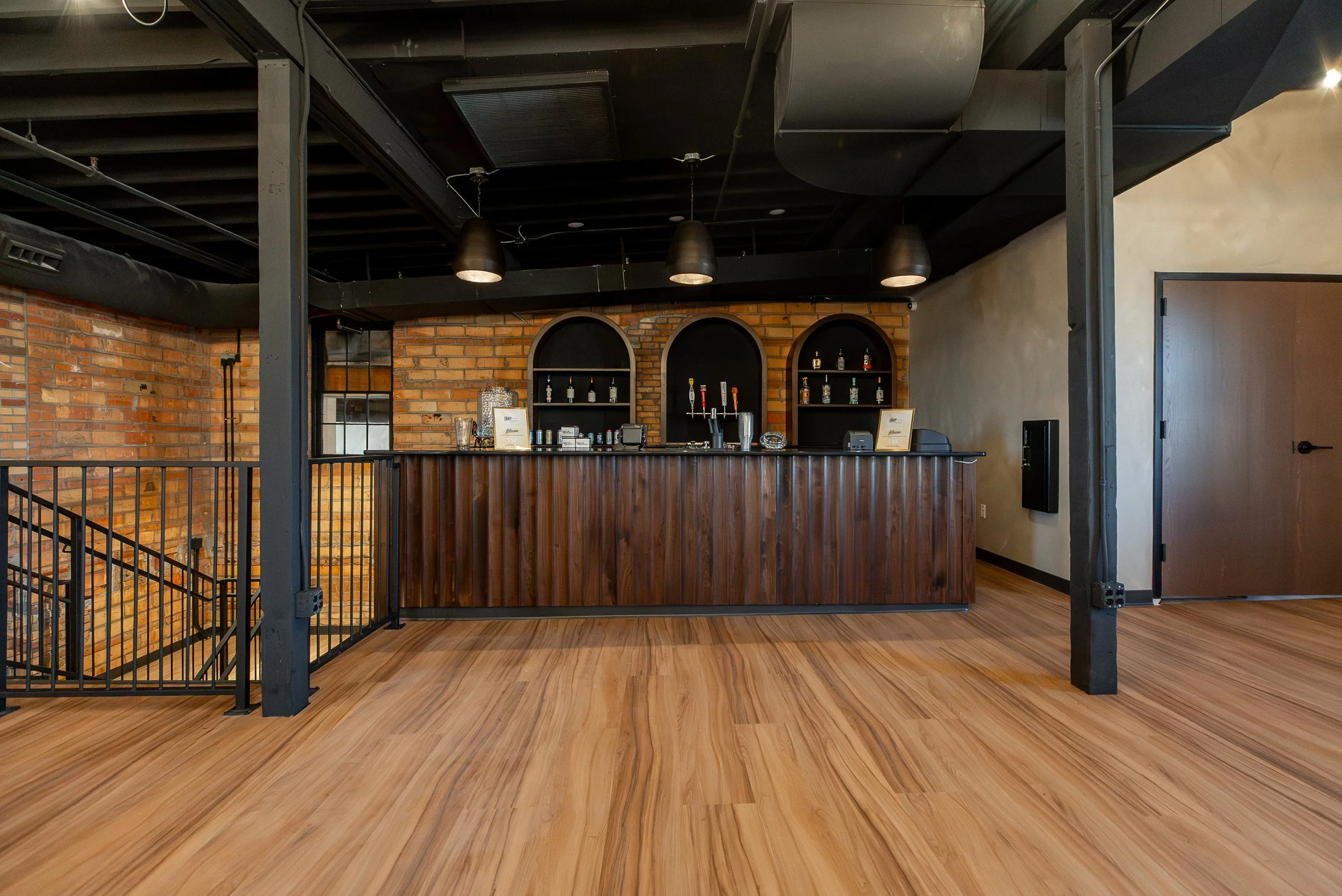 Interior of a modern bar with exposed brick wall, dark ceiling, pendant lights, and a wooden bar counter with some alcoholic bottles.