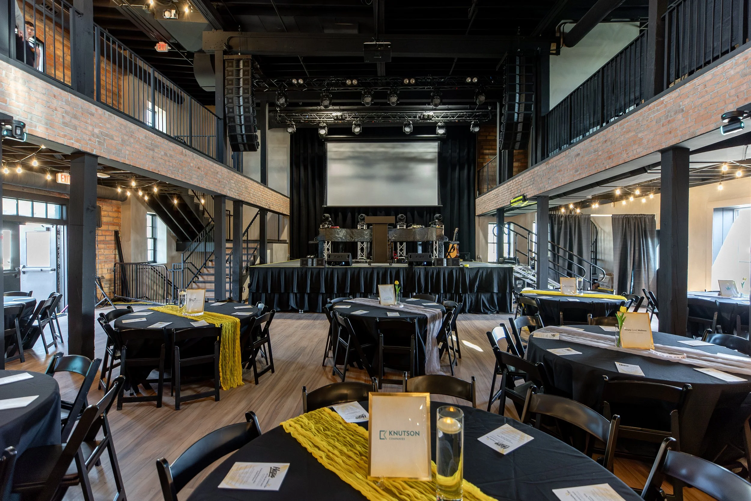 An event space with a stage and a large screen, decorated with round tables covered in black tablecloths, each with place settings and yellow runners, set up for a formal event or celebration in a modern industrial-style venue with brick walls and st