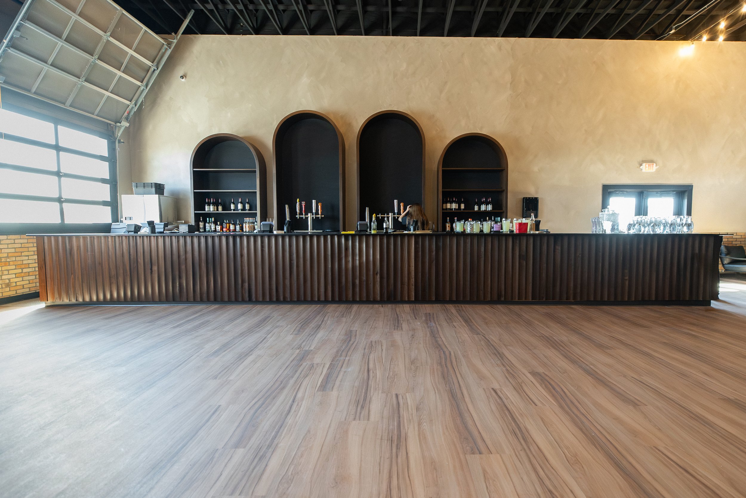 Interior of a bar with a modern design, featuring a long wooden bar counter, arched black shelving with bottles, and large windows letting in natural light.