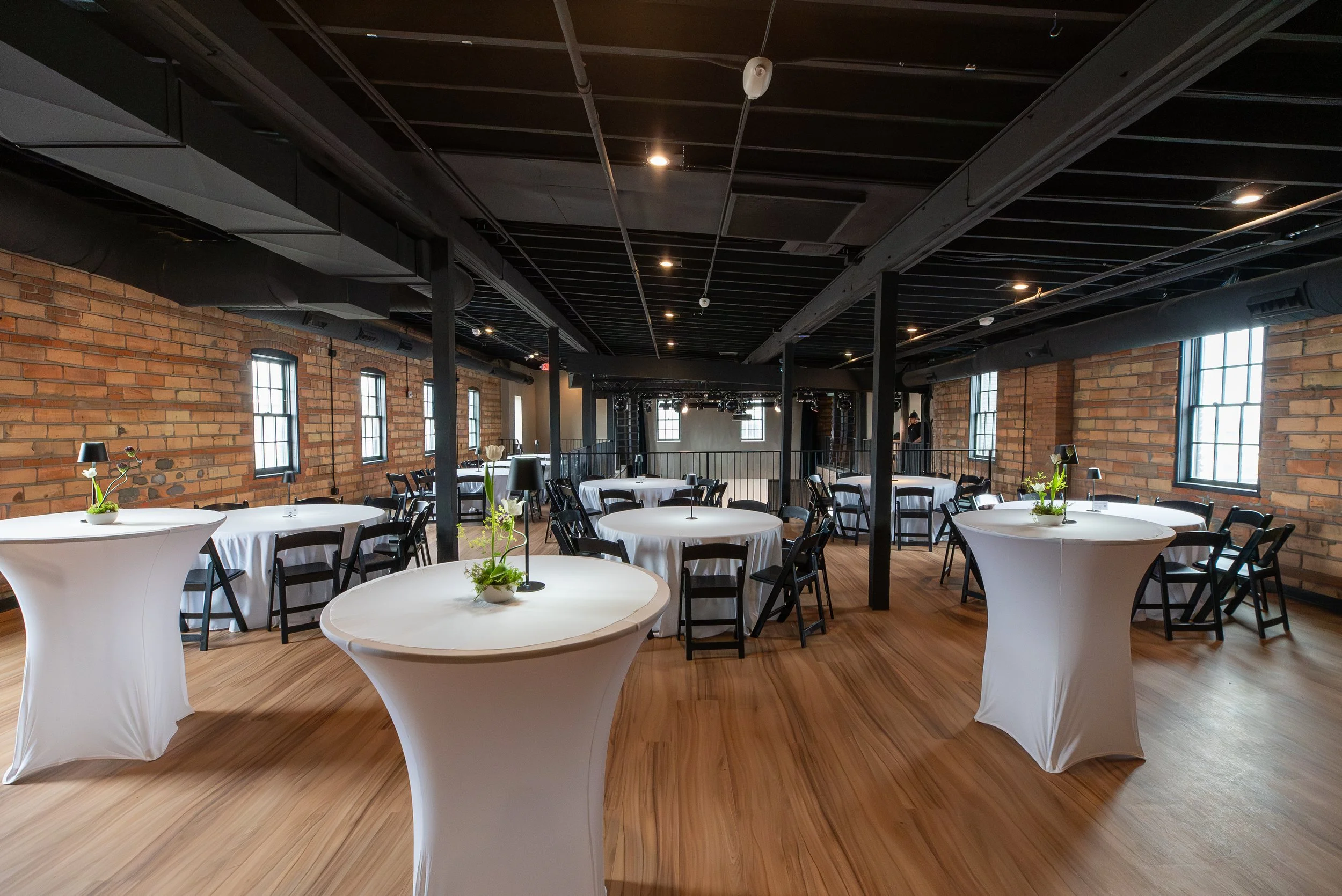 A spacious event venue with round tables covered with white tablecloths, each decorated with a small plant, set in front of exposed brick walls and large windows. The ceiling is black with visible pipes and lighting fixtures.