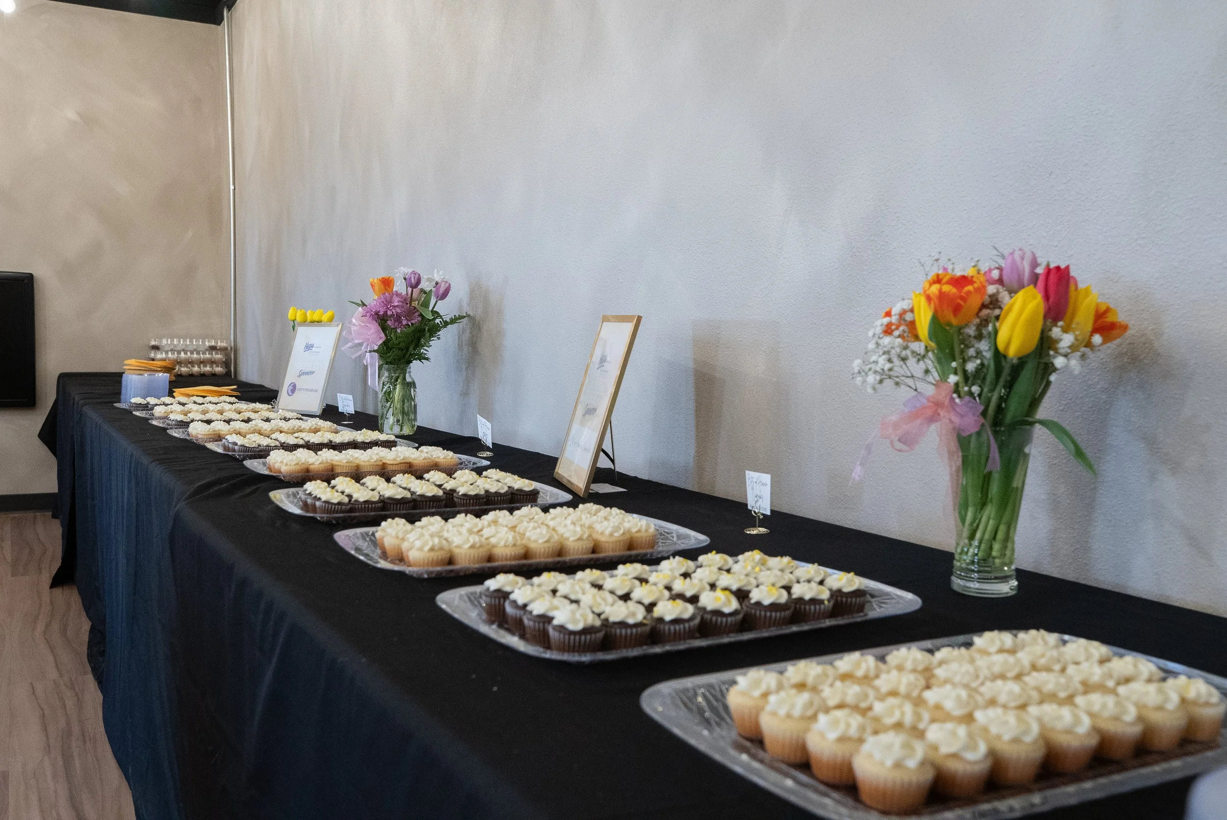 A long table covered with a black tablecloth displays various trays of cupcakes and desserts in a room with white walls. There are two floral arrangements in vases on the table, one with pink, orange, and purple flowers, and another with yellow, pink