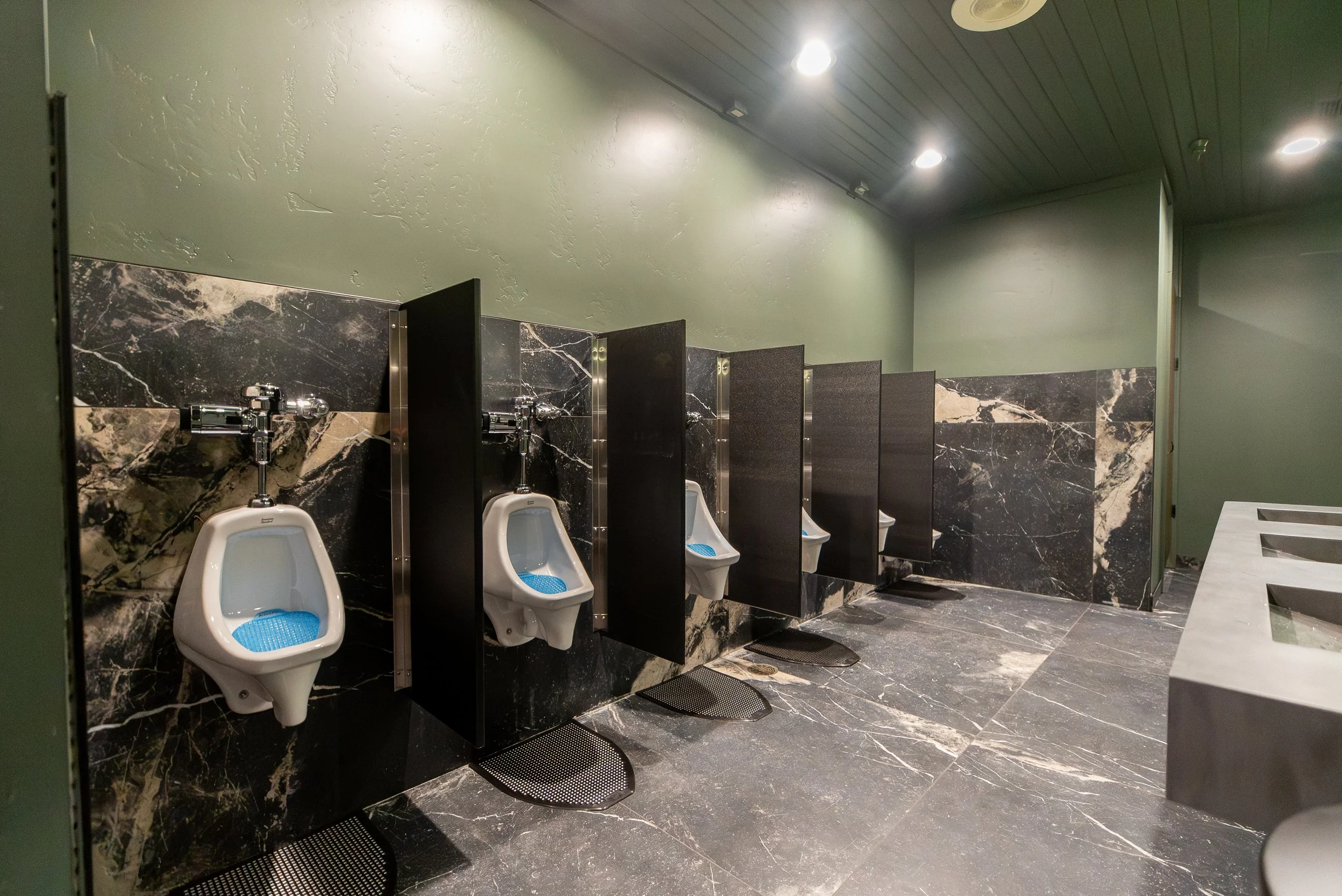 A row of four men's urinals in a public restroom with black marble walls and floor, black partitions, and a green painted wall above.