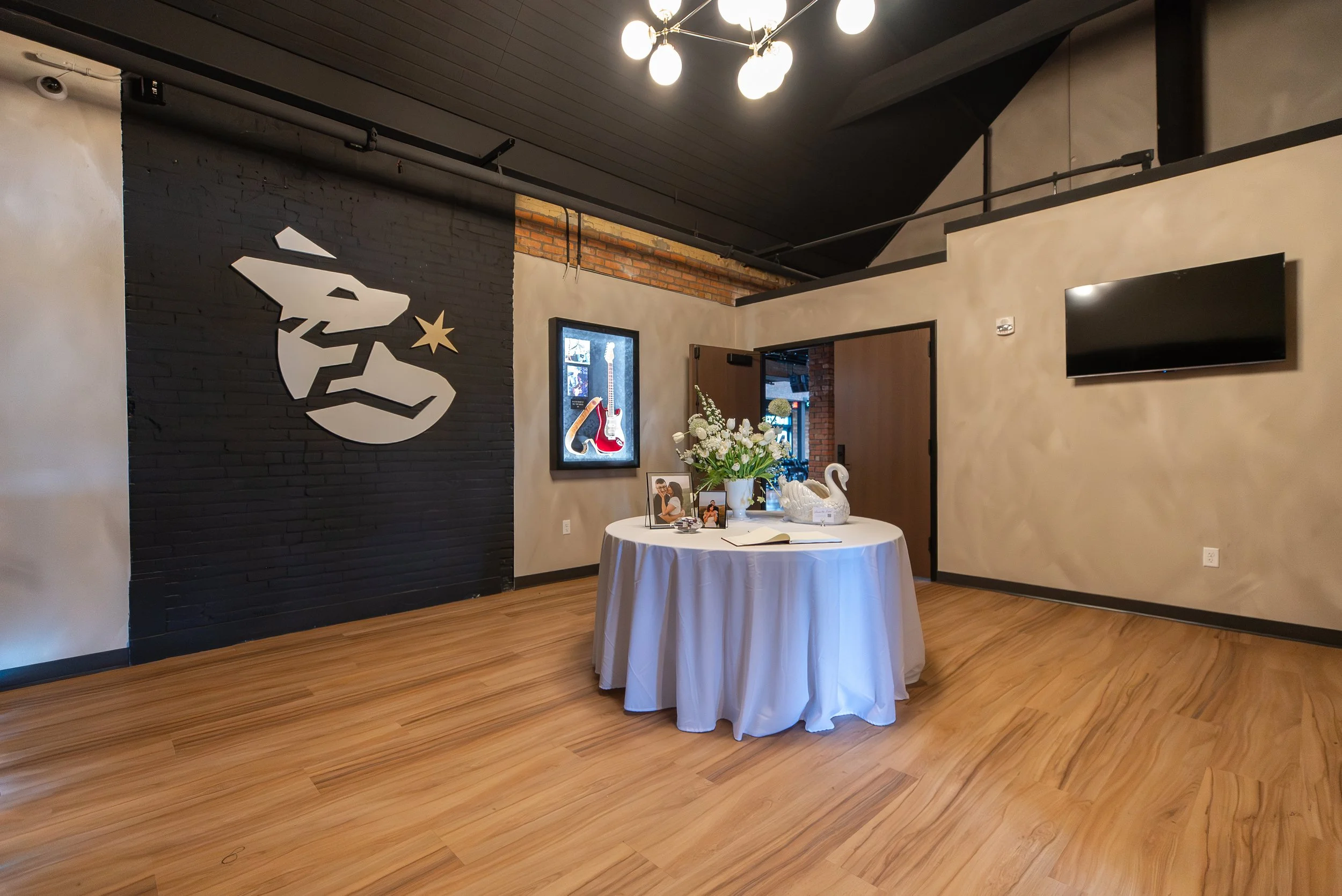 Interior of a modern room with a black wall featuring a white kangaroo silhouette and a star, a round table with a white tablecloth, floral centerpiece, framed photos, a swan sculpture, a TV on the wall, and artwork of a guitar. The ceiling has indus