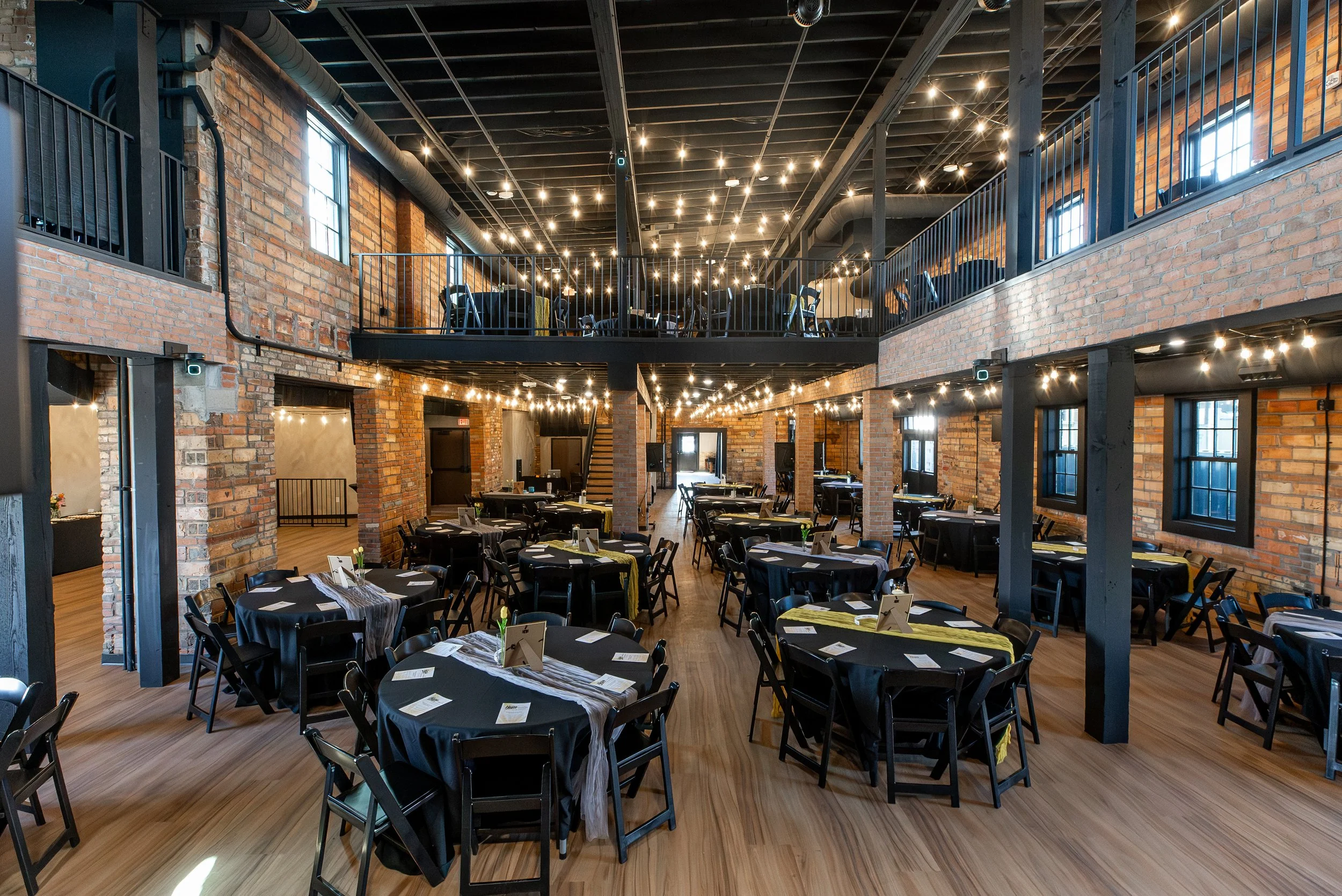 A spacious event hall with exposed brick walls, black tables with black chairs, and string lights hanging from the ceiling. There is a second-floor balcony with a black railing overlooking the main area. The room has multiple windows allowing natural