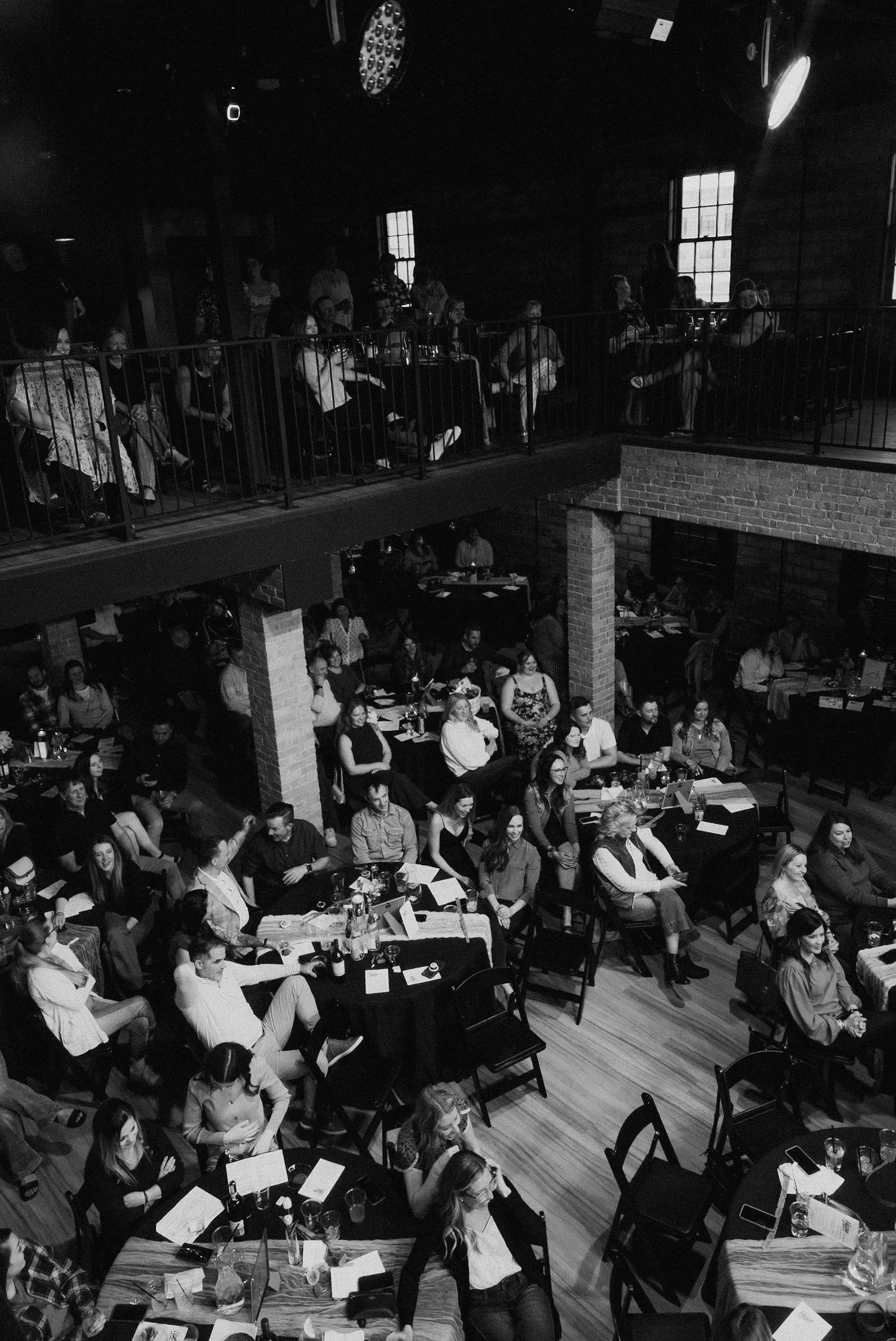 A black and white photograph of a large indoor event with many people seated at tables on a wooden floor. Guests are engaged, some holding drinks, and some standing or walking around. The venue has a two-story layout with people on the second-floor balcony observing the event. The room has wooden paneled walls and large windows, with a warm, rustic atmosphere.