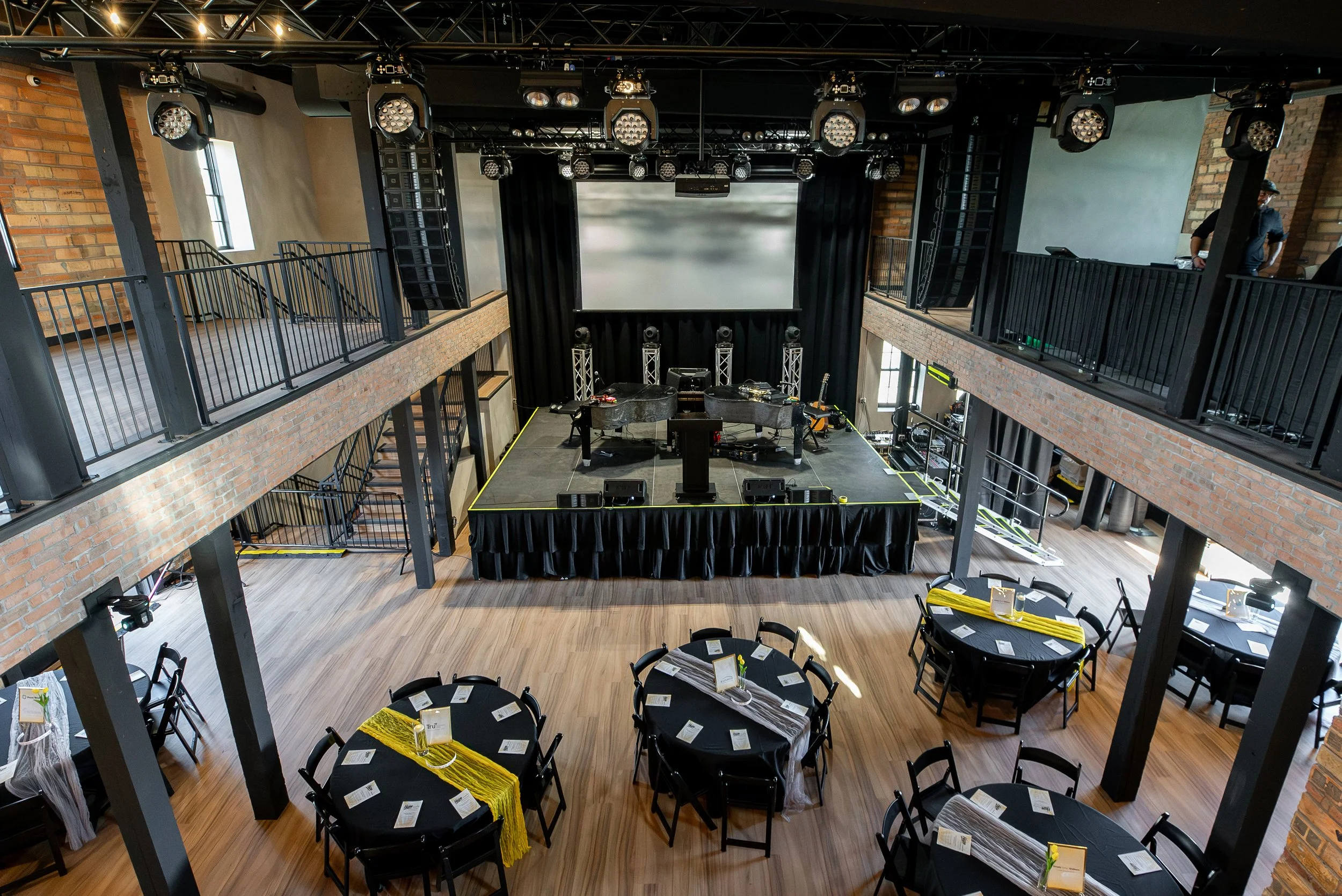 Indoor event space with round tables covered in black tablecloths and yellow runners, each set with place cards, pens, and small flower arrangements. A stage with large speakers, lighting, and a projection screen is at the front. The venue has expose