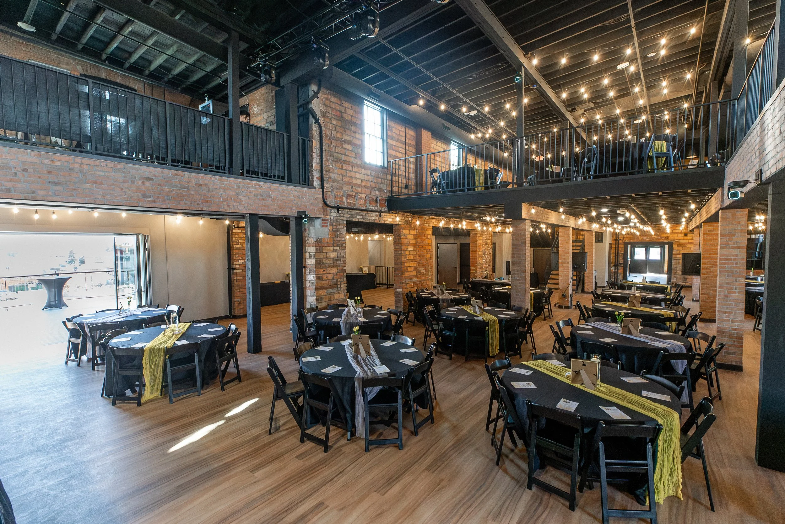 Interior of a spacious, rustic event venue with exposed brick walls and wooden floors. Round tables with black chairs are set up, each decorated with black tablecloths and yellow table runners. The ceiling features string lights, creating a warm ambi