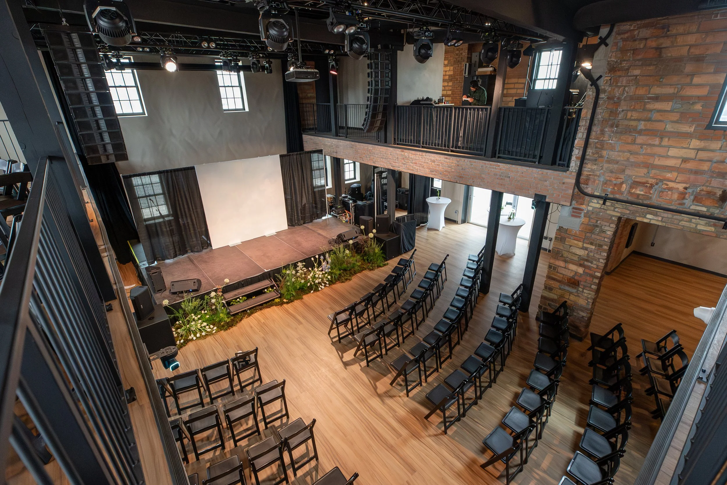 An indoor event space with wooden floors, black chairs arranged in rows, a stage with flowers, large windows, black curtains, and a white projection screen. There is a balcony with a DJ and audio equipment, and brick walls.