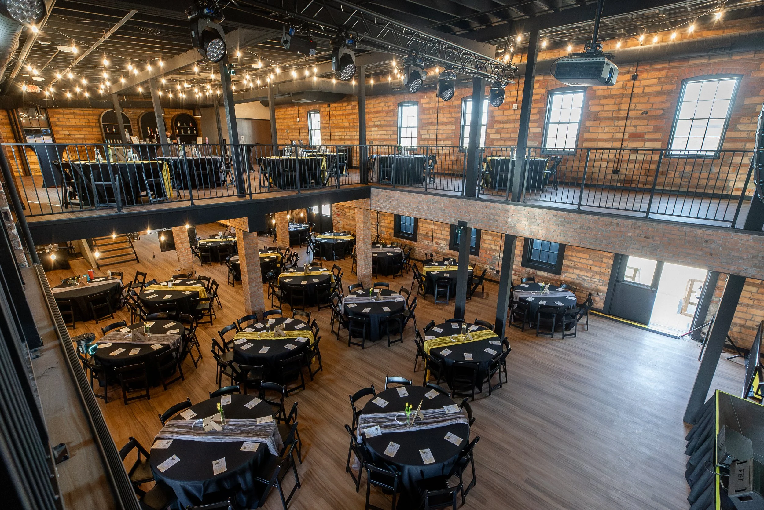 Interior of a rustic-style event space with exposed brick walls, wooden flooring, and black tables with yellow accents set for a meal. The space has large windows, string lights overhead, and a second-floor balcony with additional tables and seating,