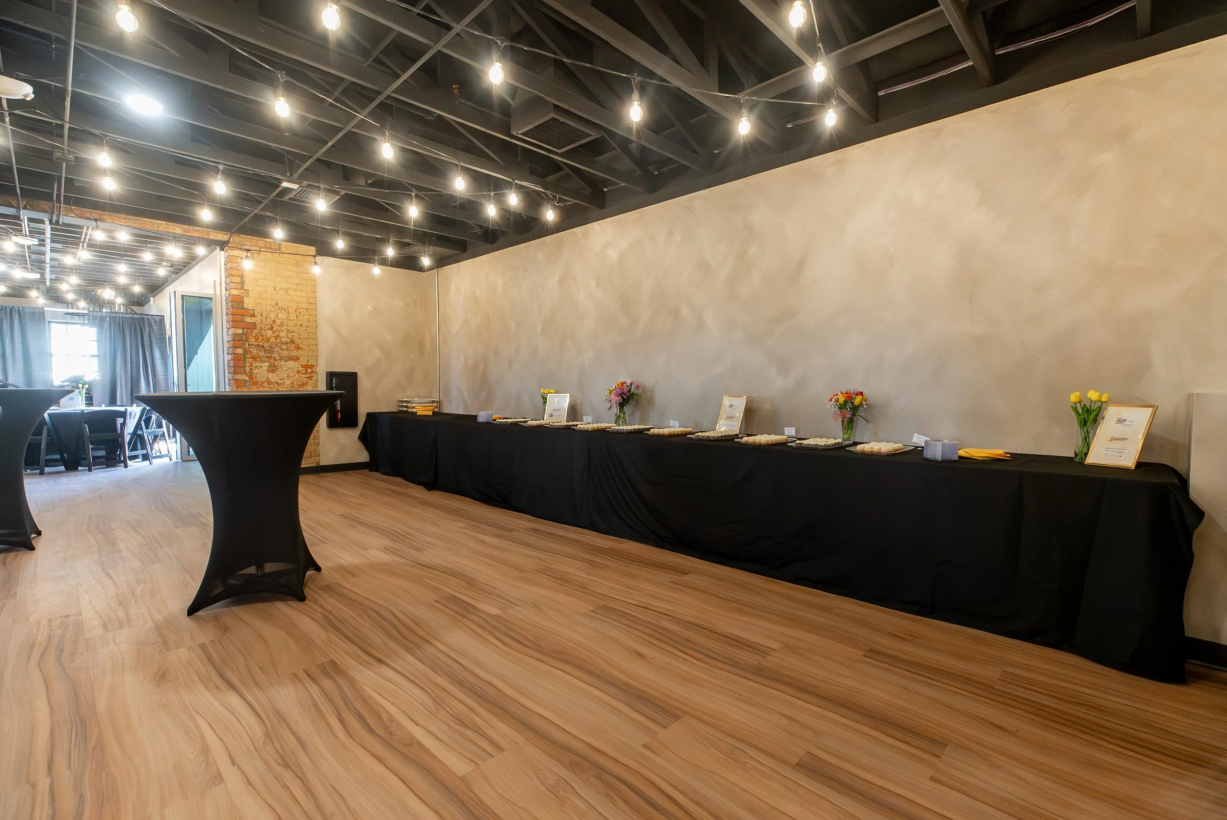 Interior of event space with string lights on black ceiling beams, a long black table with flowers and signs, and a small round table with a black cloth.