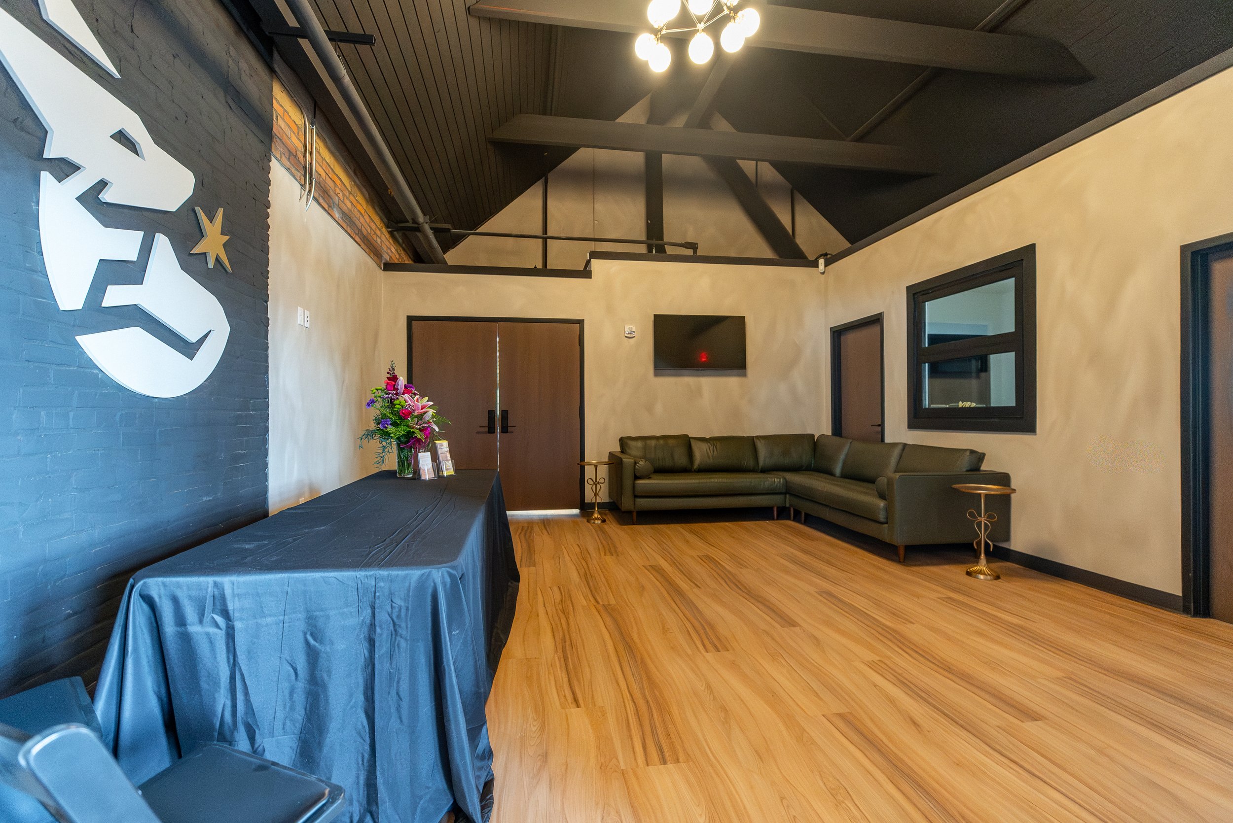 Interior of a modern lounge or waiting area with a black brick accent wall featuring a white and gold deer silhouette, a table with a floral arrangement, a long dark sofa, and a wall-mounted TV. The room has wood flooring, black window frames, and a 