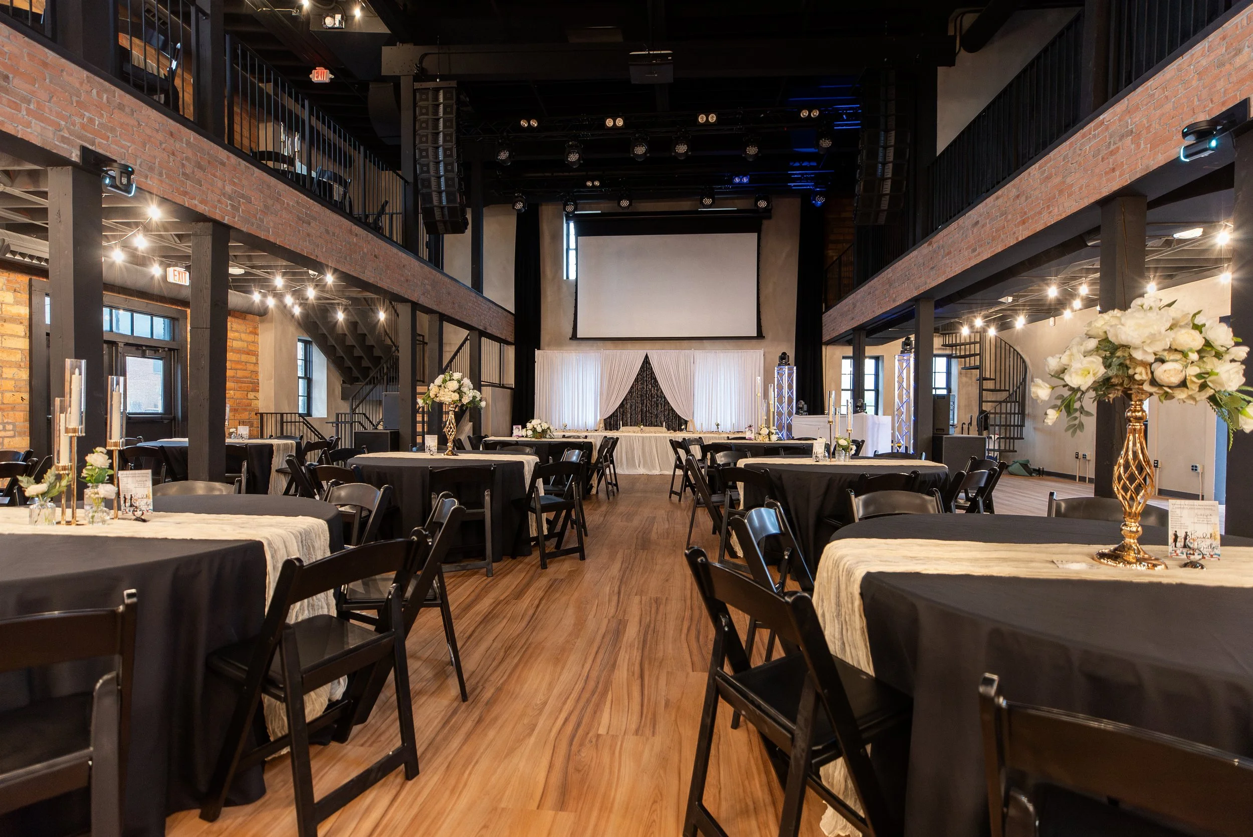 Interior of a wedding reception hall with round tables covered in black and white tablecloths, floral centerpieces, and a stage with a white curtain backdrop. Elevated balcony with black railings runs along both sides, exposed brick walls, and string lights hanging from the ceiling.