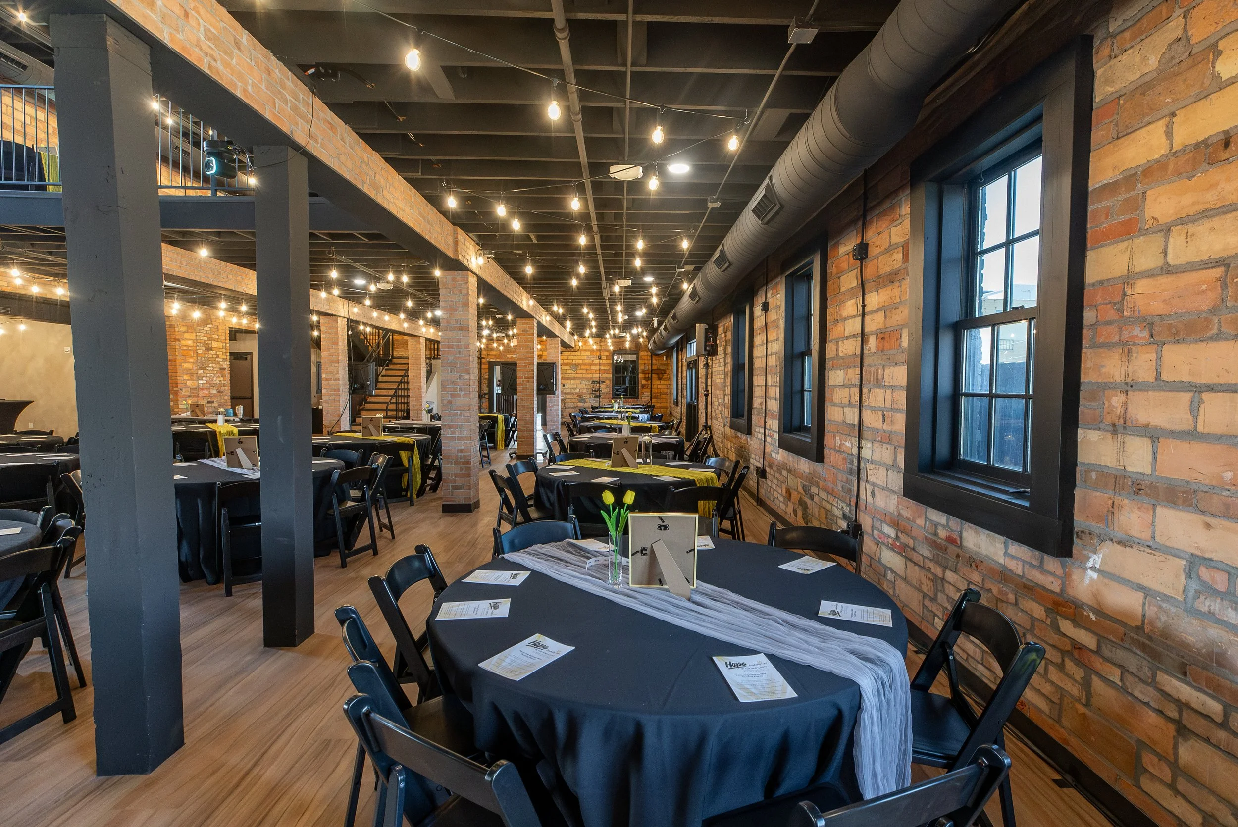 Interior of a rustic-style event space with brick walls, string lights hanging from the black-painted ceiling, and tables set up with black tablecloths and placemats for a formal dinner or gathering.
