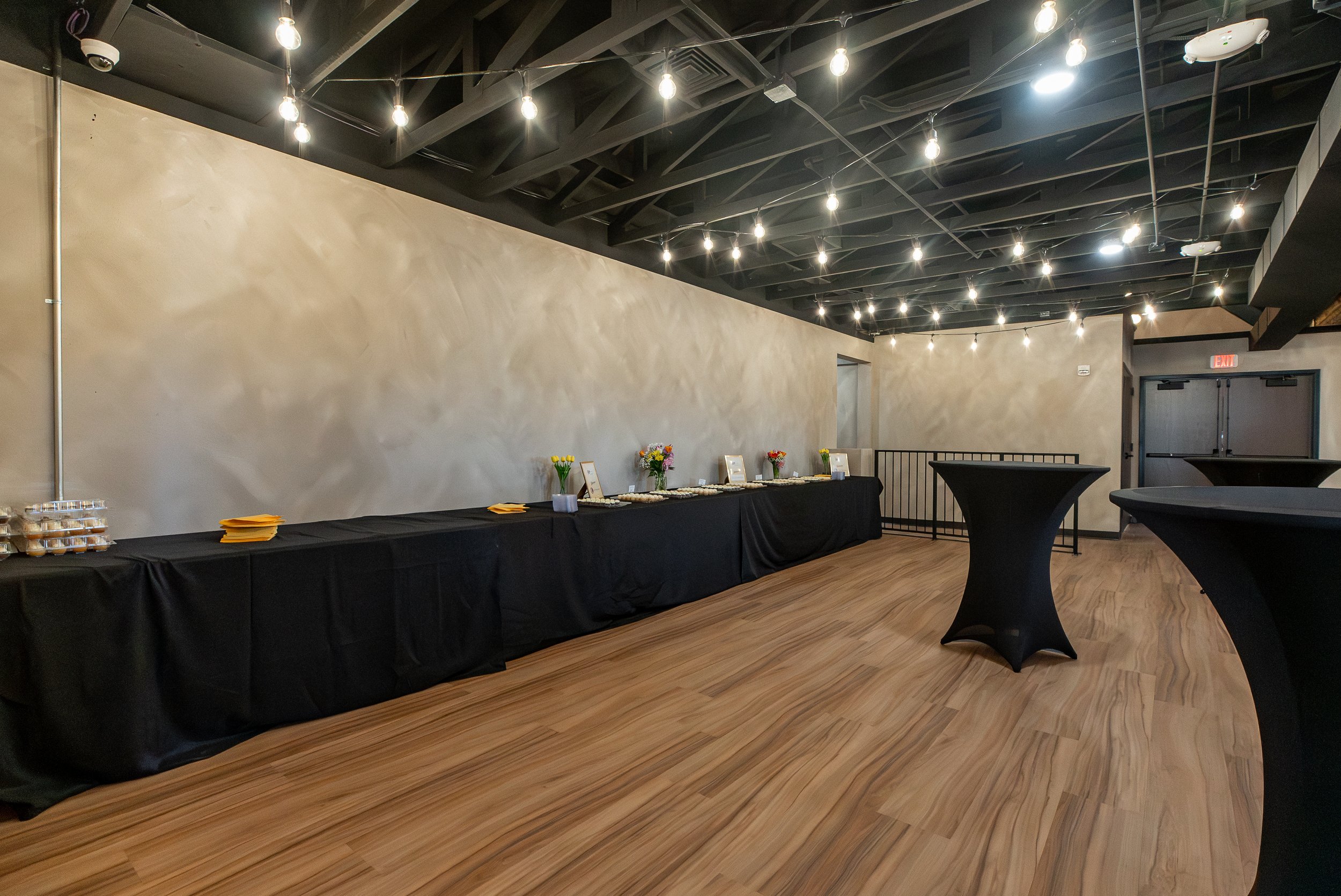 An indoor event space with a black ceiling featuring exposed beams and hanging string lights, beige textured walls, and a hardwood floor. There are black tables with flower arrangements and a long table with desserts and yellow napkins, along with a 