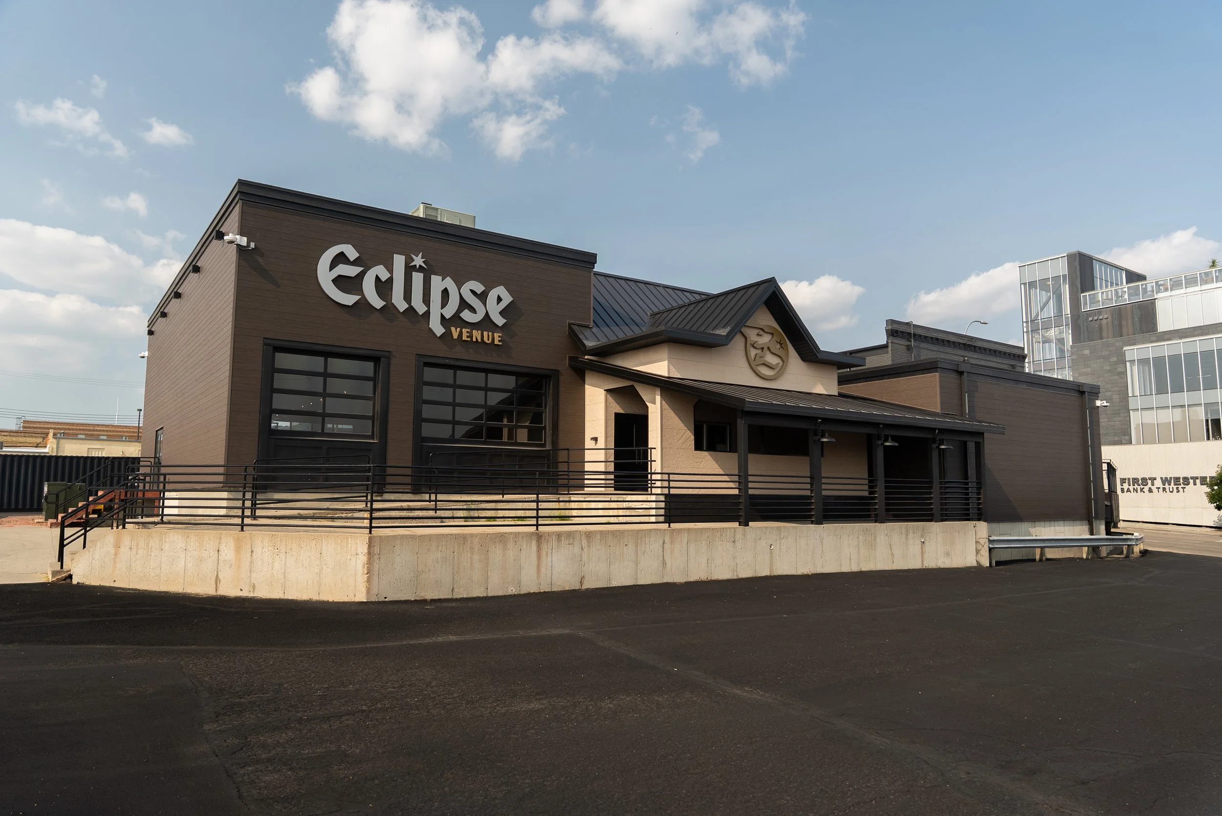Exterior of a venue called Eclipse Venue with large windows, a black roof, and decorative masks on the front wall. The sky is partly cloudy, and the pavement is empty.