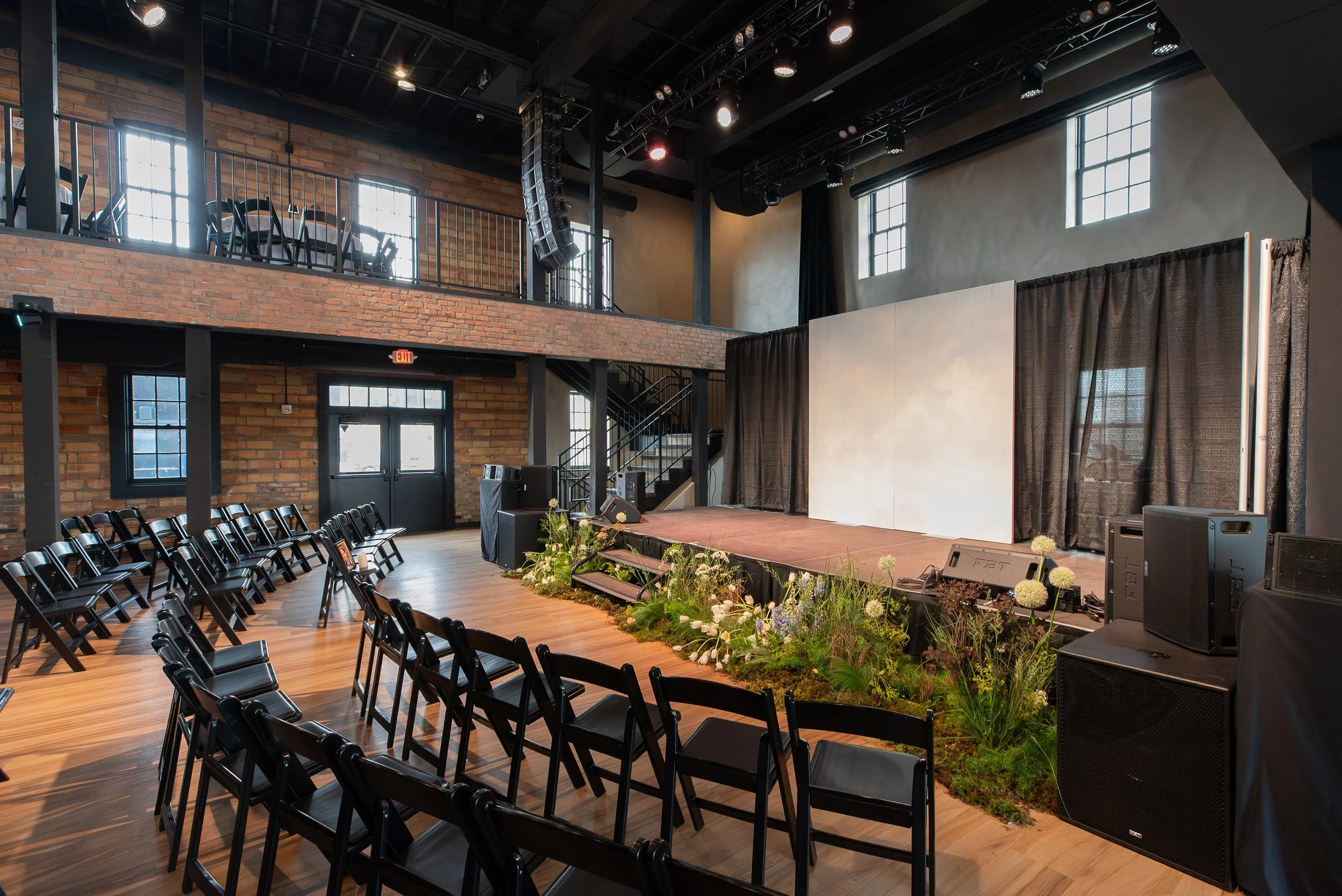 Empty stage with floral decorations, chairs arranged in rows, and a scenic backdrop inside a large hall with brick walls, multiple windows, and balcony area.