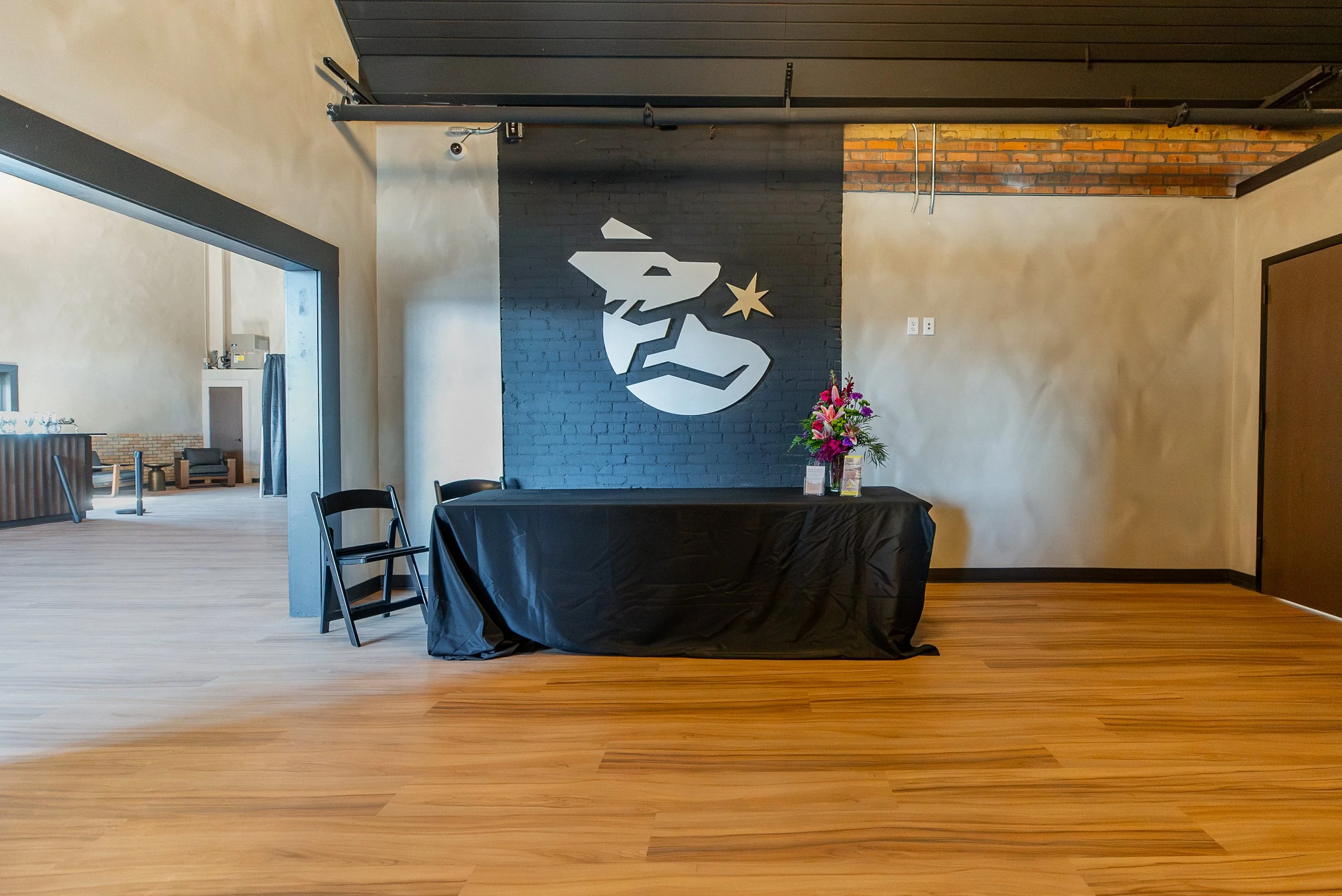 Interior space with a black accent wall featuring a white angular bird and star logo, a table with a flower arrangement, chairs, an open doorway revealing a seating area, and a ceiling with exposed pipes.