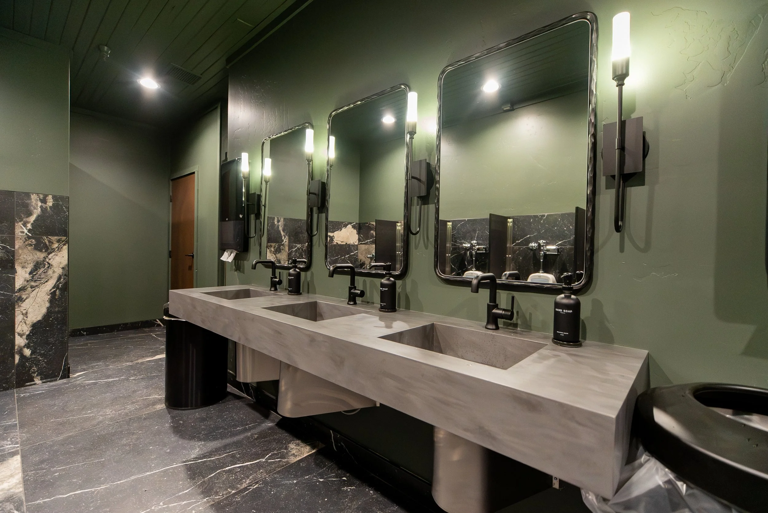 Public restroom with three sinks, mirrors, hand soap dispensers, and wall-mounted hand dryers, with dark marble flooring and green-painted walls.