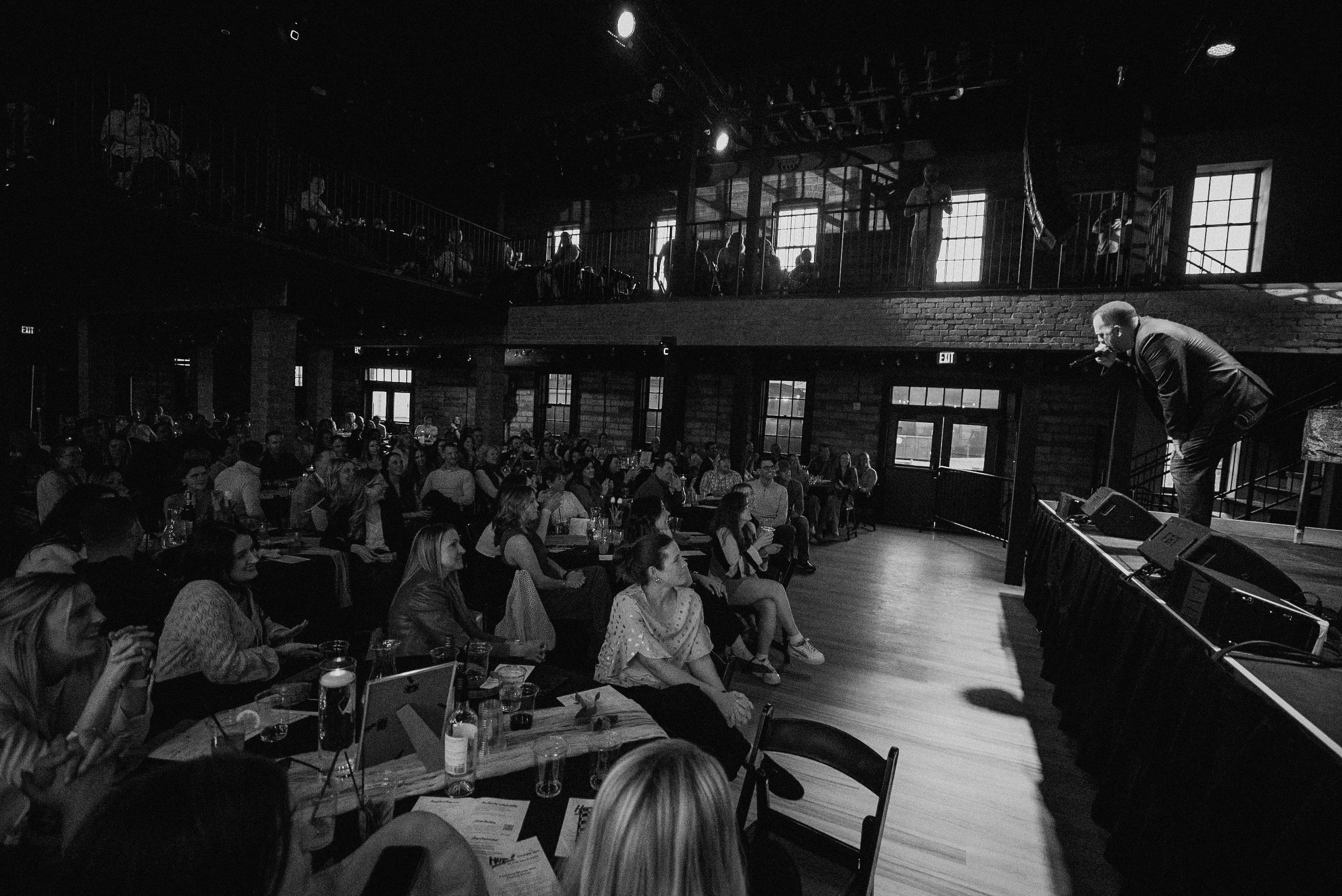 A man performs on stage with a microphone in hand, addressing a seated audience in a dark, industrial-style venue with large brick walls, tall windows, and a balcony area above.