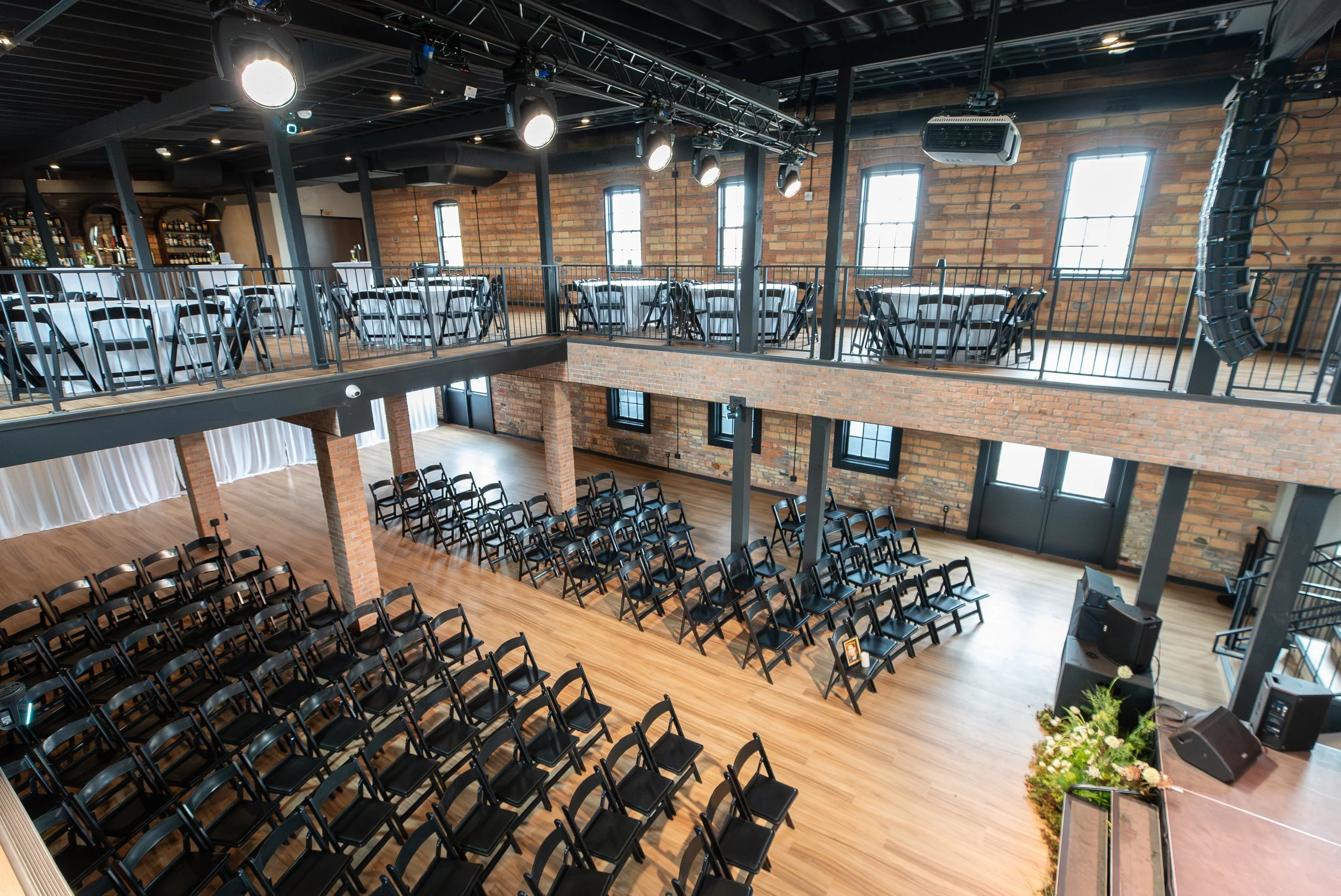 An indoor event space with a brick wall interior, large windows, and balconies with tables and chairs set up for a gathering, with stage lighting and sound equipment.