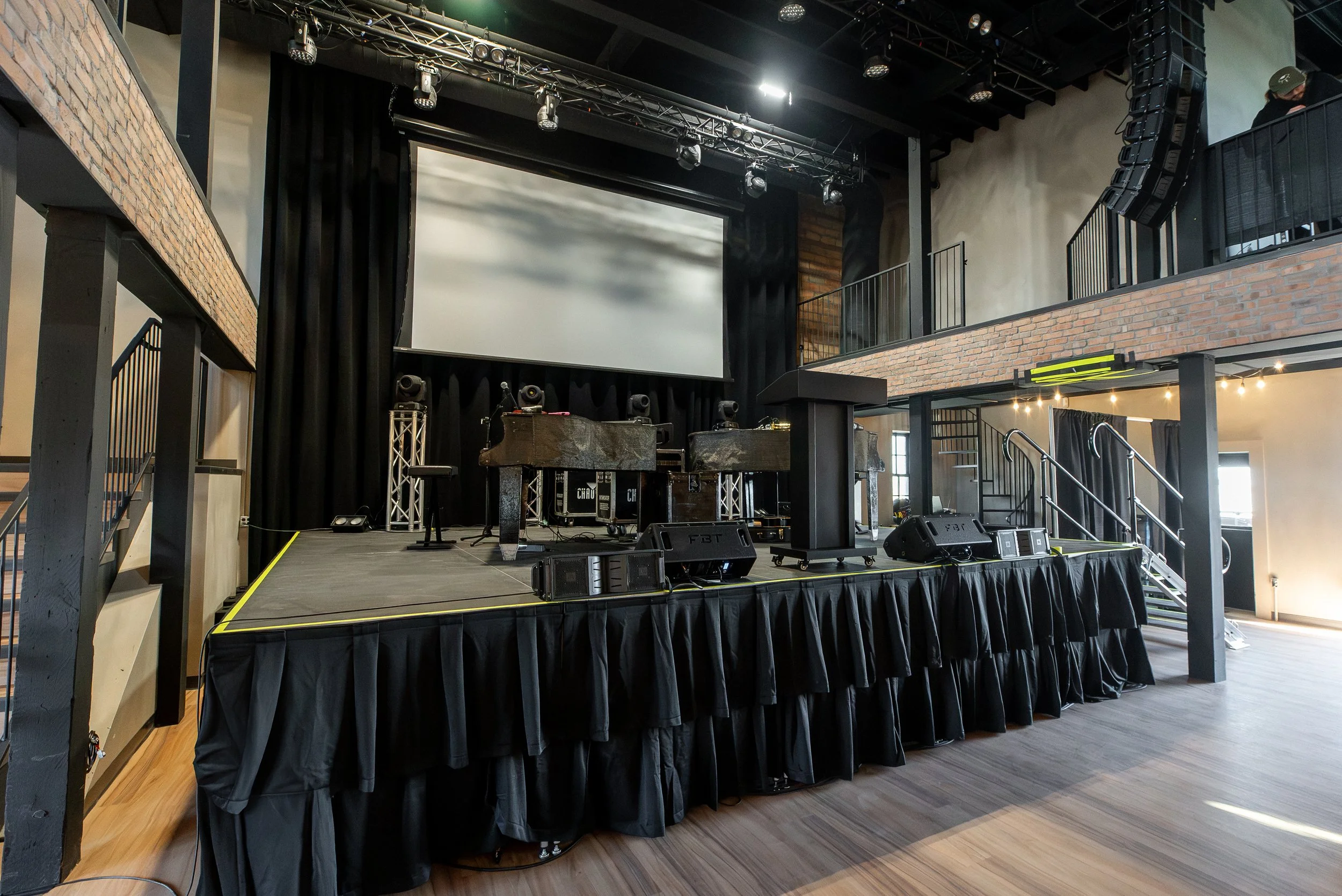 A stage set up with sound and lighting equipment, a large screen, black curtains, and an upper-level balcony with a person working on sound equipment. The stage has black tablecloth-covered tables, speakers, and a microphone.