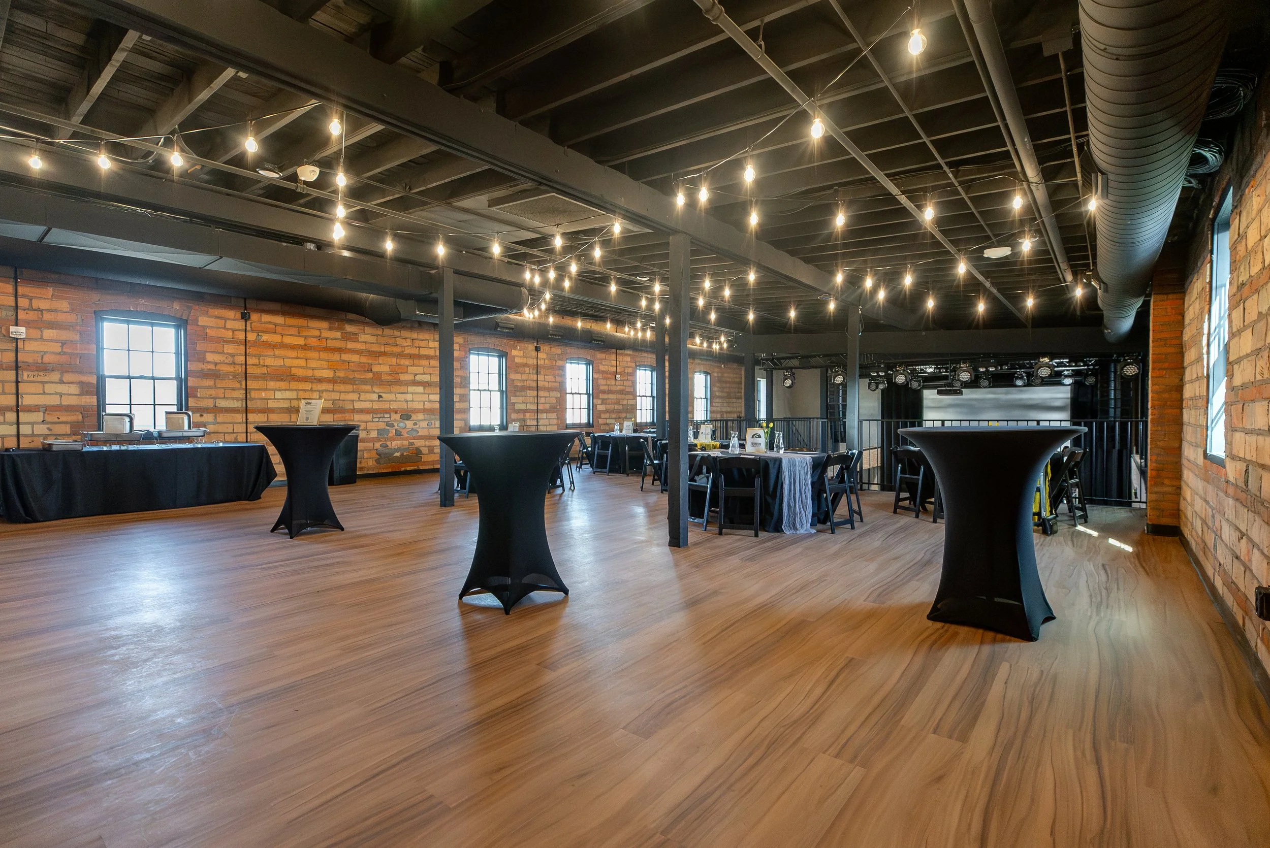 Event space with high ceilings, string lights, brick walls, wooden floors, and black high-top tables with tablecloths, set up for a gathering or celebration.