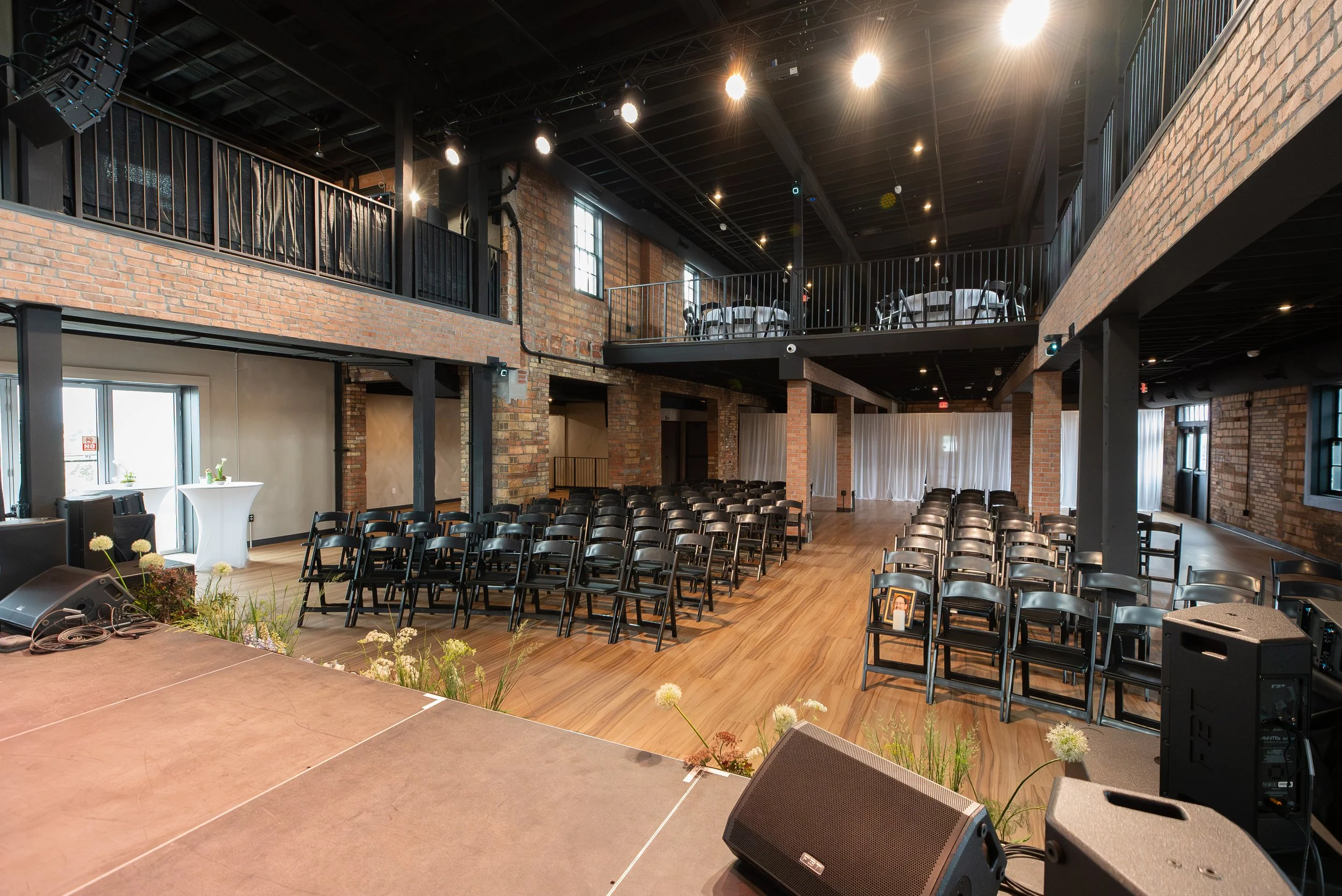 Empty event space with rows of black chairs facing a stage, exposed brick walls, large windows, and a balcony area with tables and chairs, illuminated by ceiling lights.