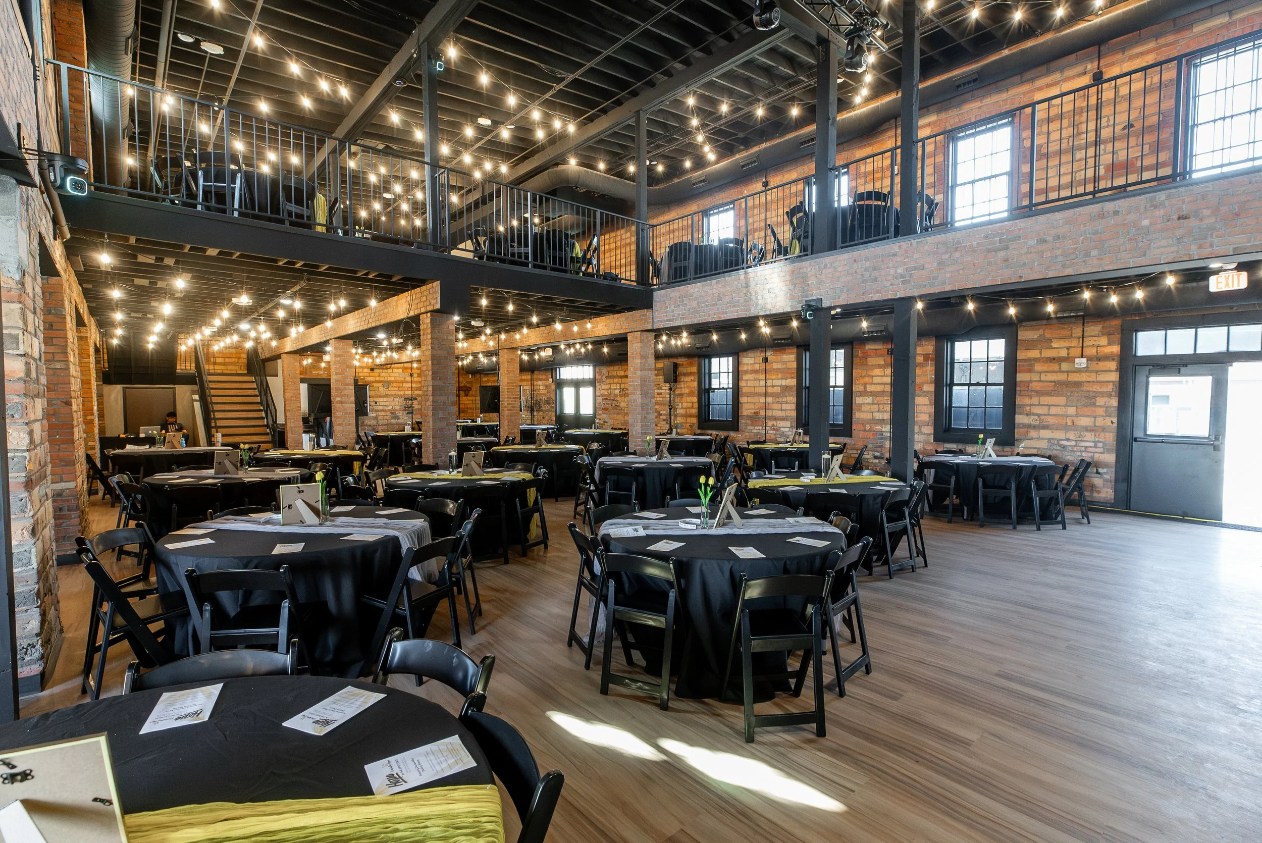 Interior of a modern restaurant with black tables and chairs, exposed brick walls, and string lights hanging from the ceiling. There is a balcony area with additional seating.