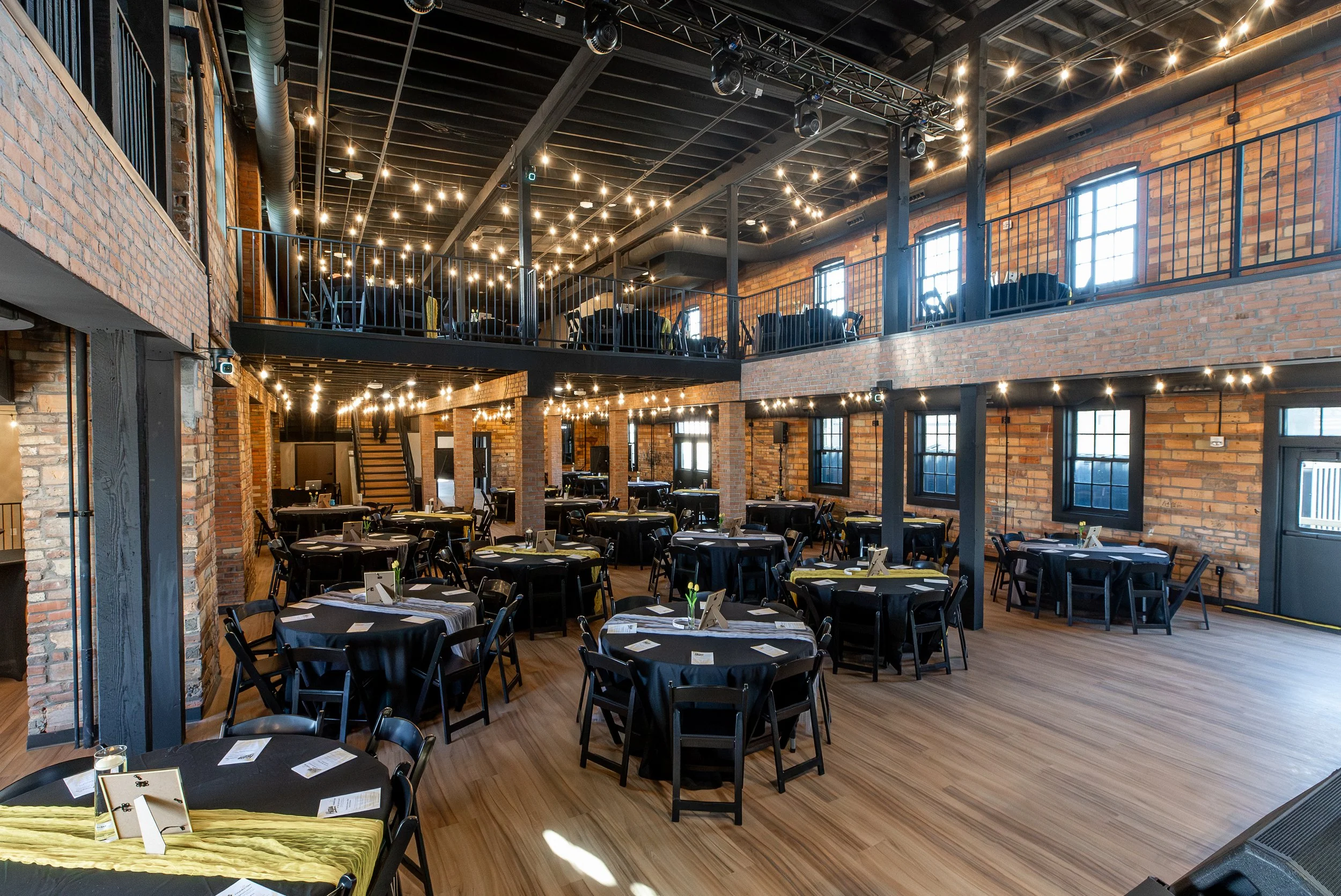 The image shows a spacious, industrial-style event venue with exposed brick walls, large windows, and a black ceiling with string lights. There are round tables with black chairs, set up for a formal event or celebration.