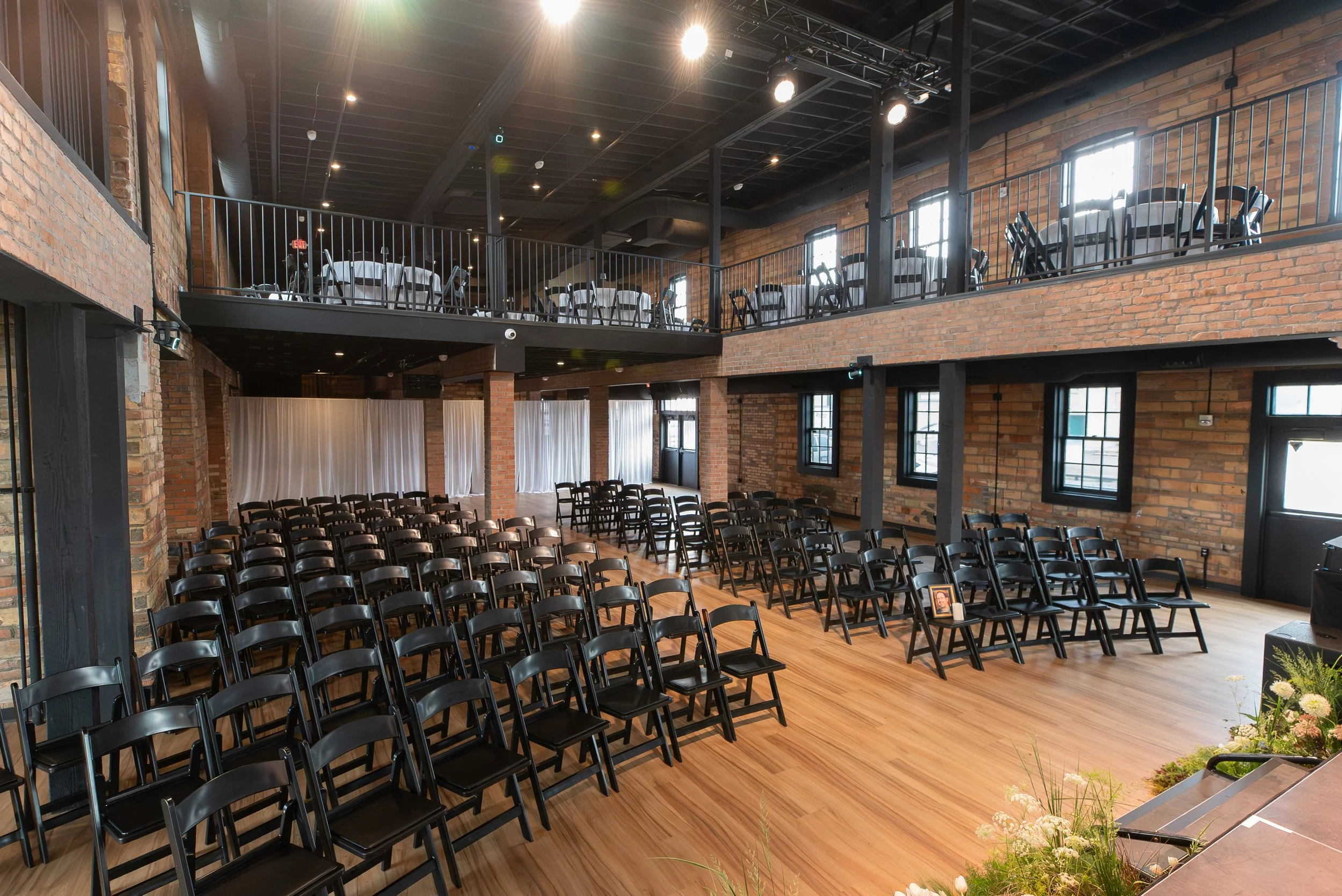 An indoor event space with rows of black chairs facing a stage. The space has exposed brick walls, large windows, and a wooden floor. There is a balcony with tables and chairs above the main area, and decorative floral arrangements in the foreground.