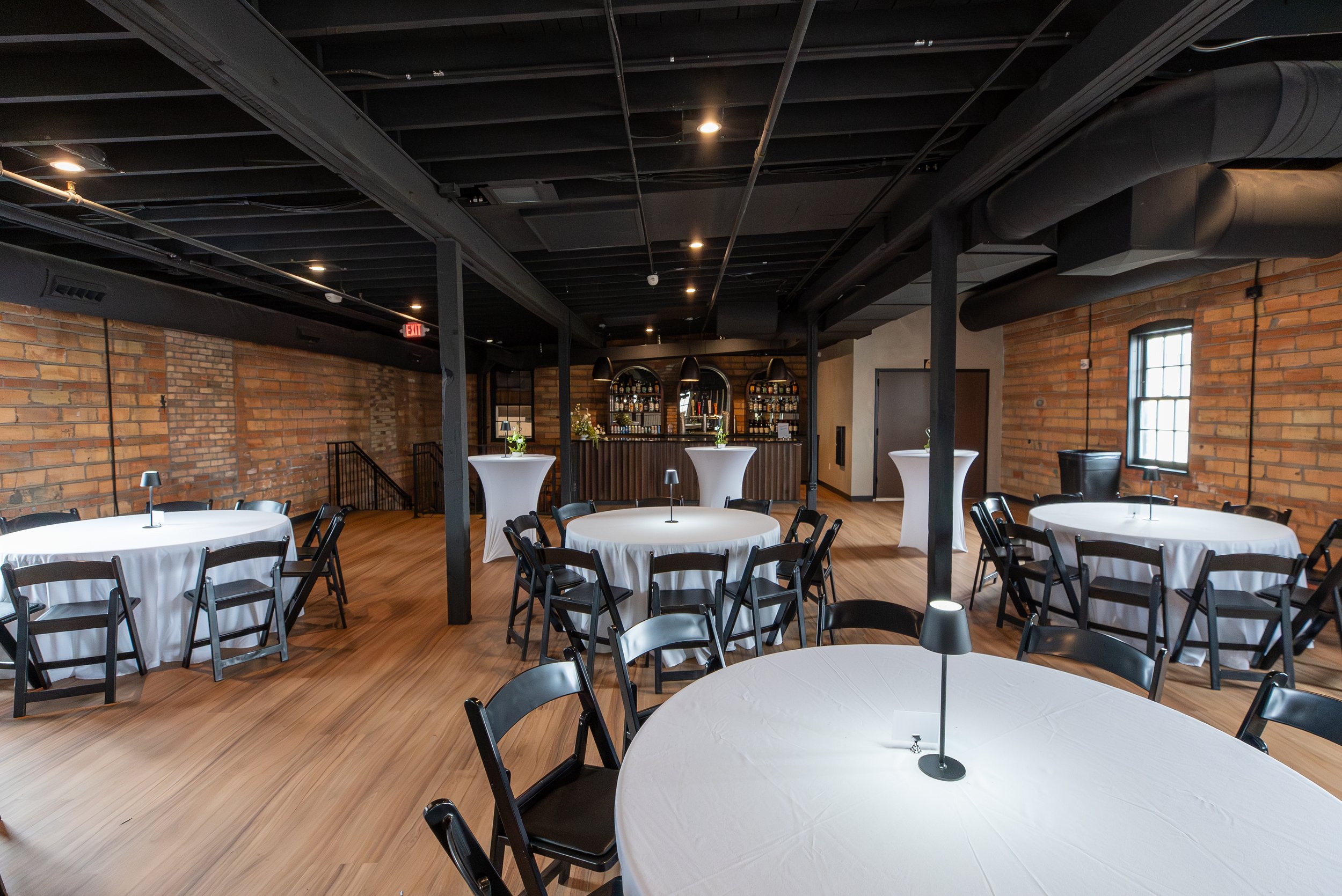 Blank banquet or event space with round tables covered in white tablecloths and black chairs, exposed brick walls, large windows, and a bar area in the background.