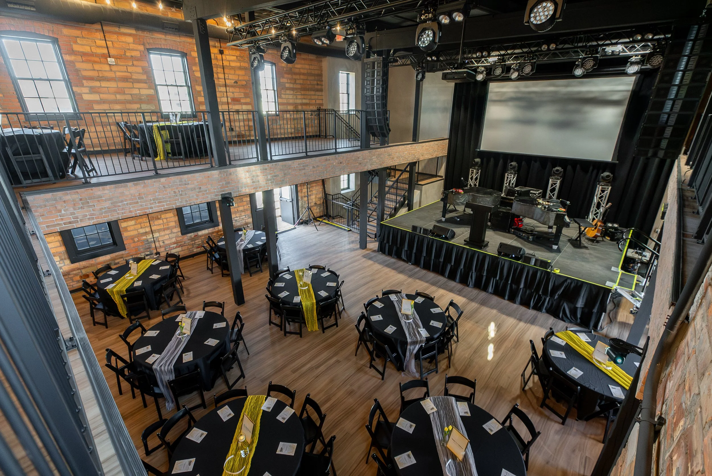 Event space with round tables decorated with black tablecloths and yellow runners, arranged for a gathering. A large stage with a screen, musical instruments, and lighting equipment is visible. The area has brick walls and large windows, with an uppe