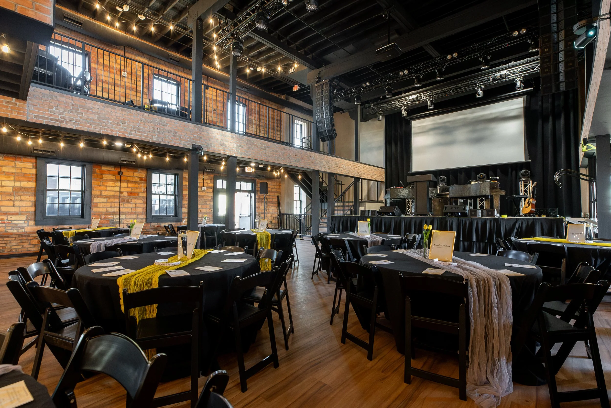 An indoor event space or auditorium with a stage, black curtains, and musical instruments set up, including a guitar and speakers. There are round tables with black tablecloths and yellow table runners, and the space features brick walls, large windo