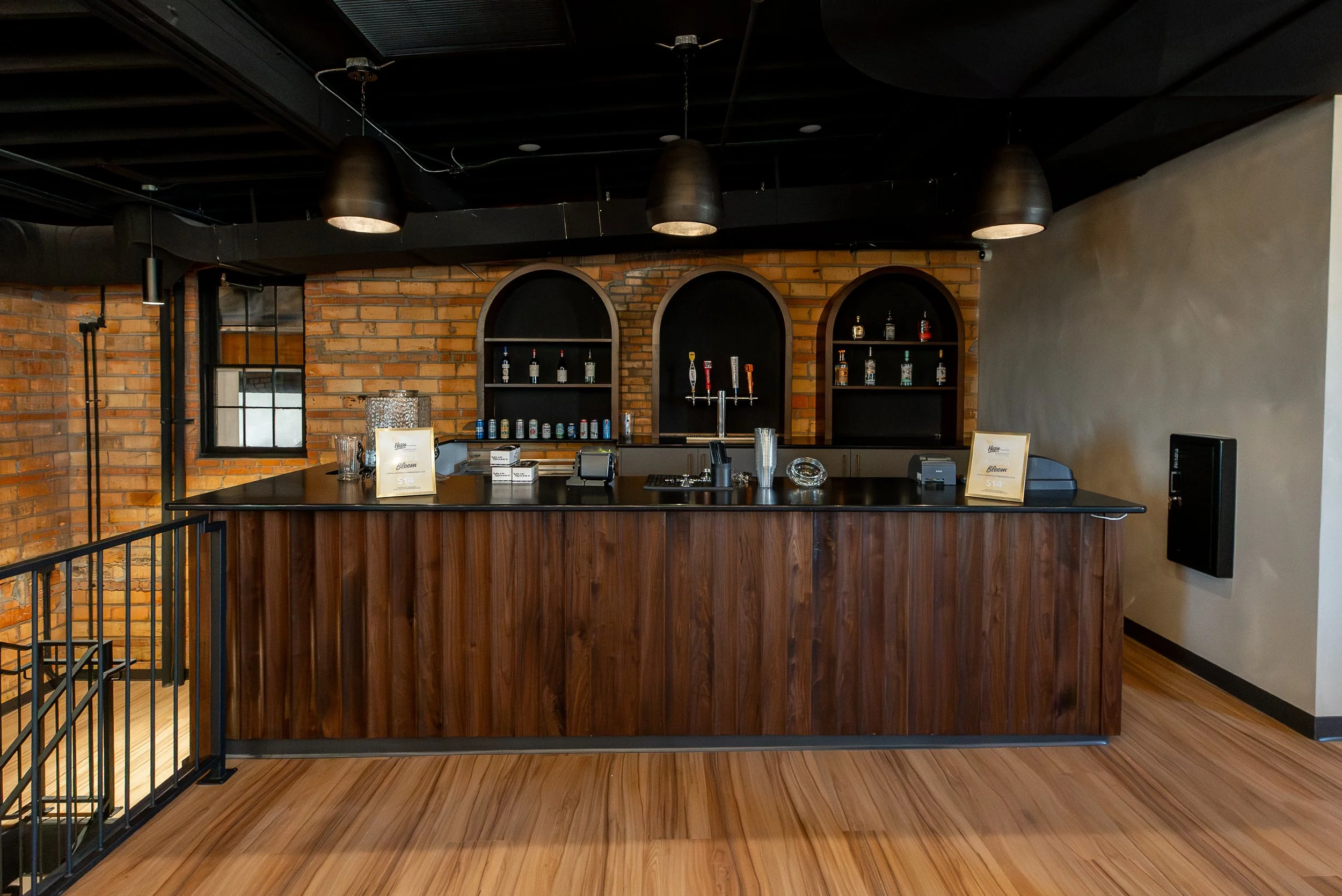 Bar counter with three shelves behind it, stocked with liquor bottles and cans, in a bar with exposed brick walls and black ceiling with hanging lighting fixtures.