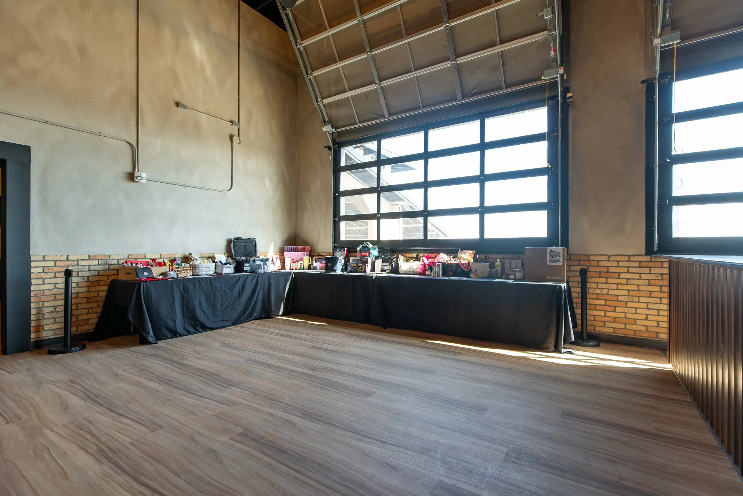 An indoor event space with a high angled ceiling and large windows, featuring a long table covered with a black tablecloth and various items, including baskets and boxes, against a backdrop of brick and wooden panel walls.