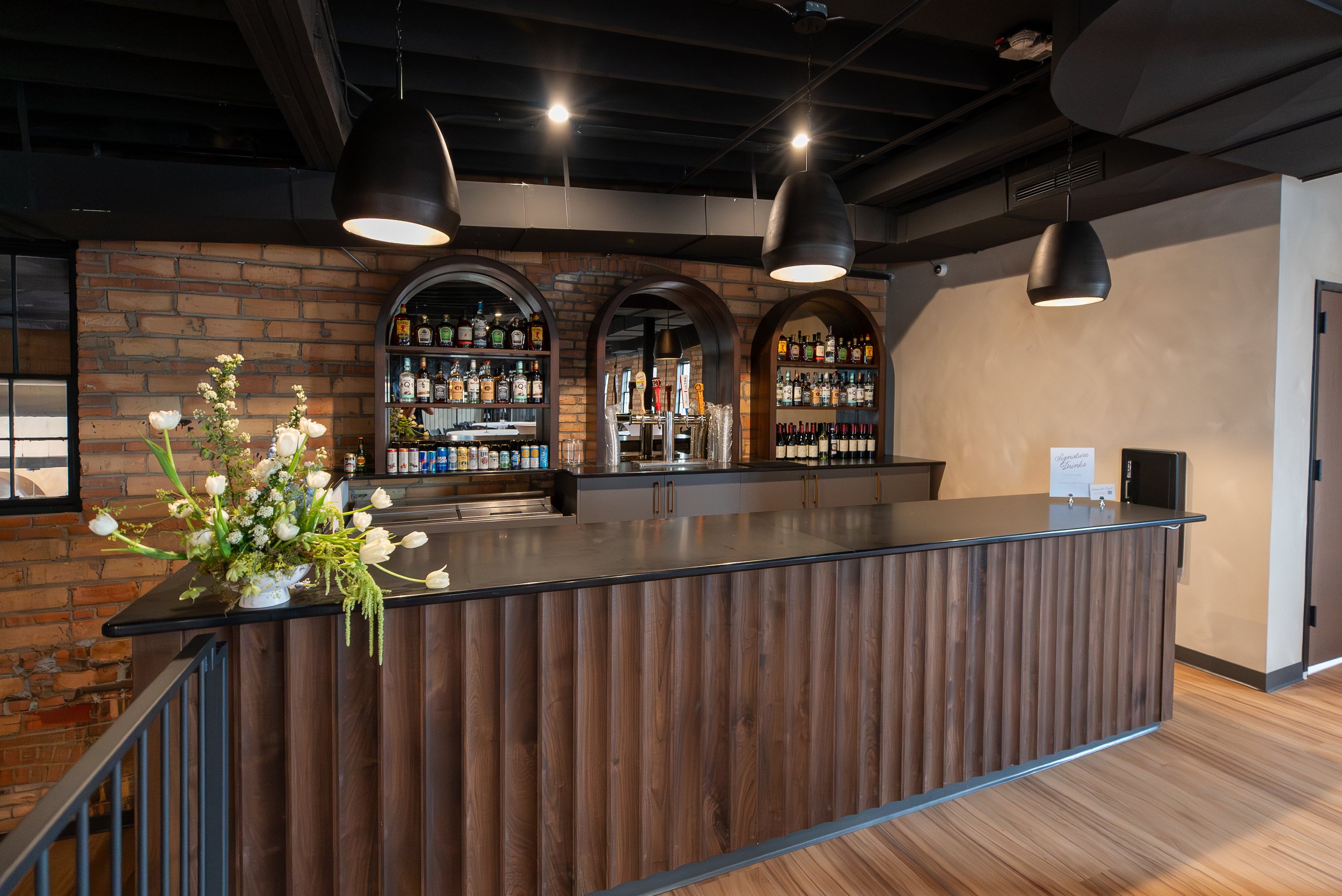 Interior of a bar area with a counter, decorated with a flower arrangement, brick wall, and shelves with various bottles of alcohol, illuminated by hanging black pendant lights.