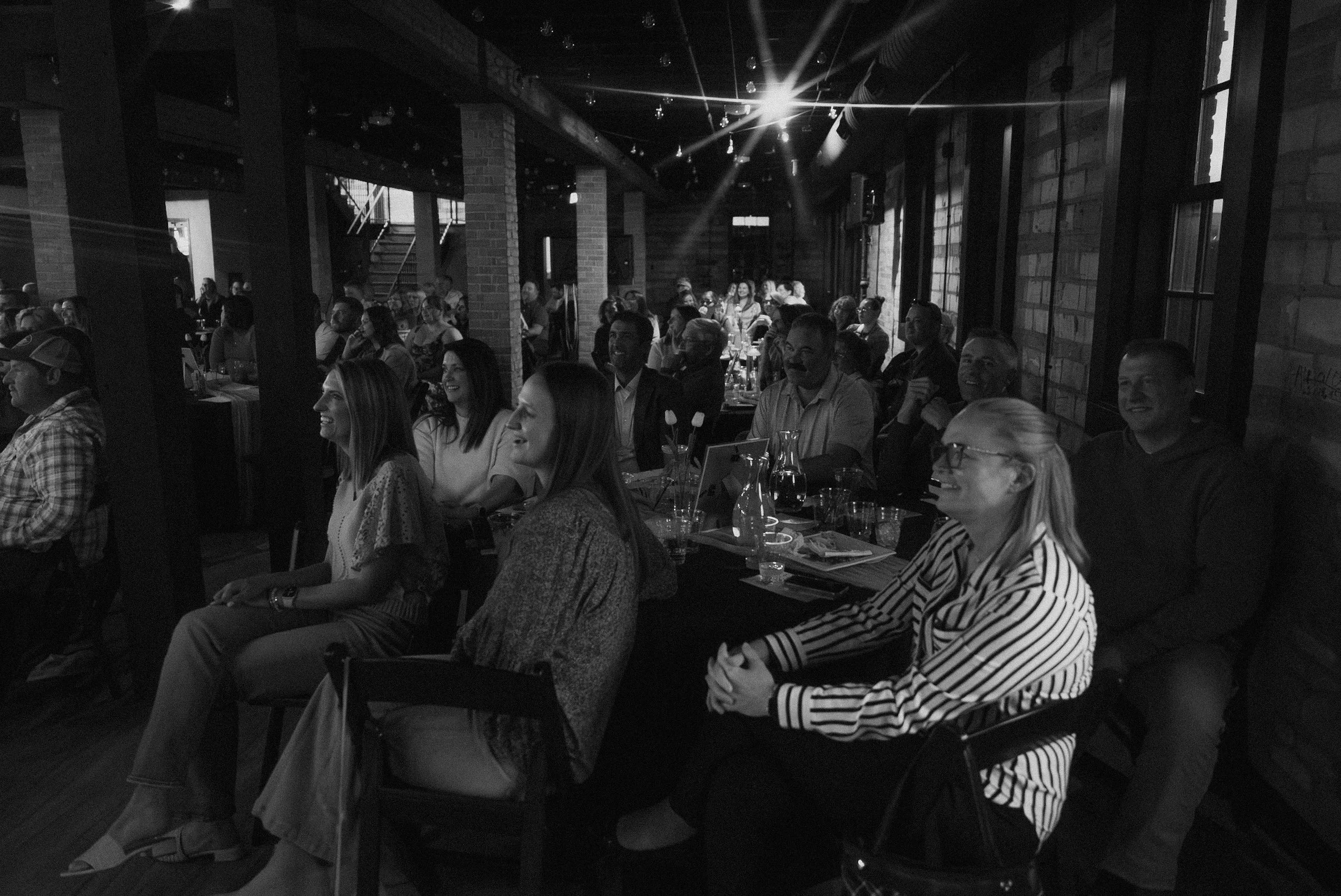 A group of people sitting at tables in a dimly lit venue, possibly attending a concert or event, with some smiling and others engaged in conversation.