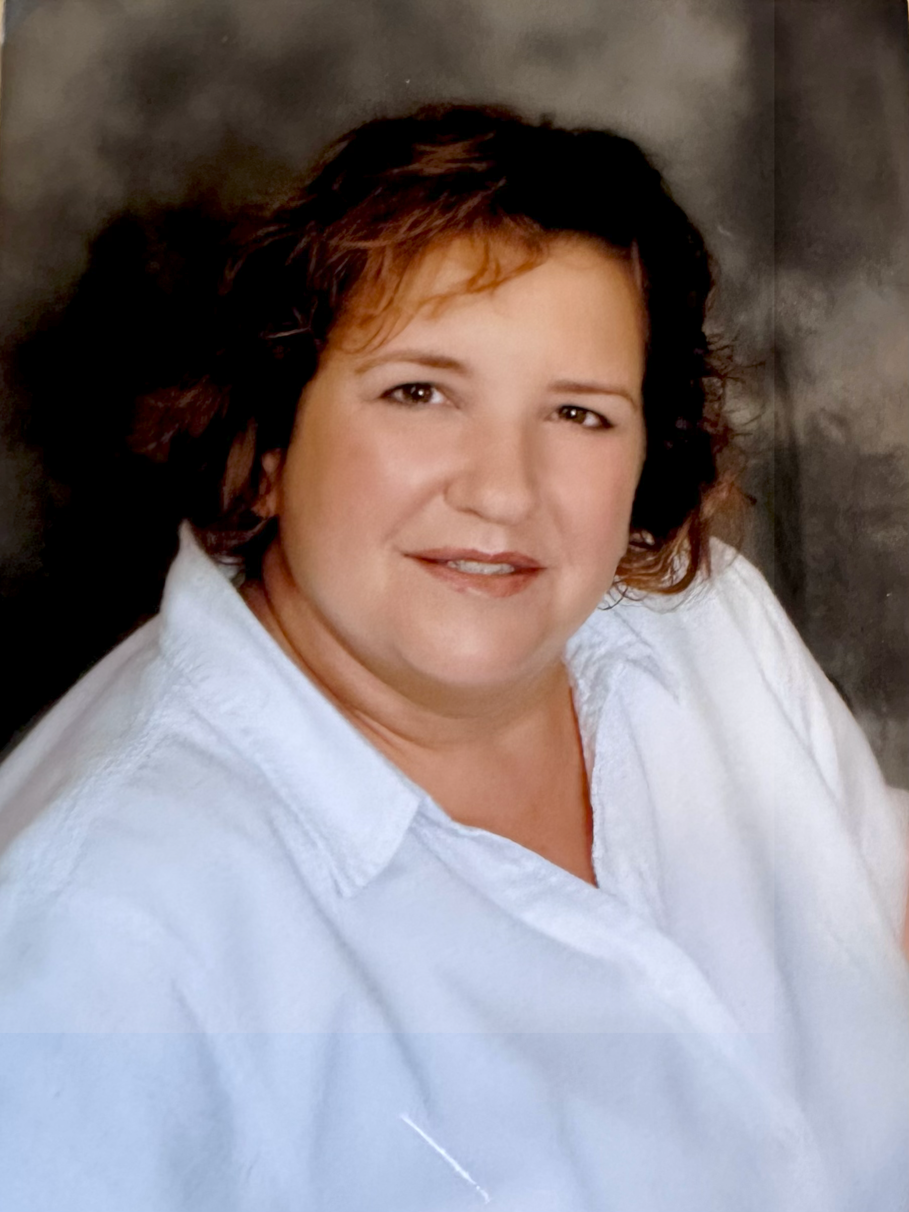 A woman with short brown hair wearing a white collared shirt, smiling softly at the camera against a dark background.