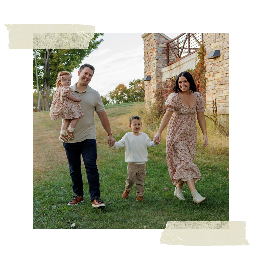 Family walking outdoors on grass, holding hands, smiling, with trees and stone building in background