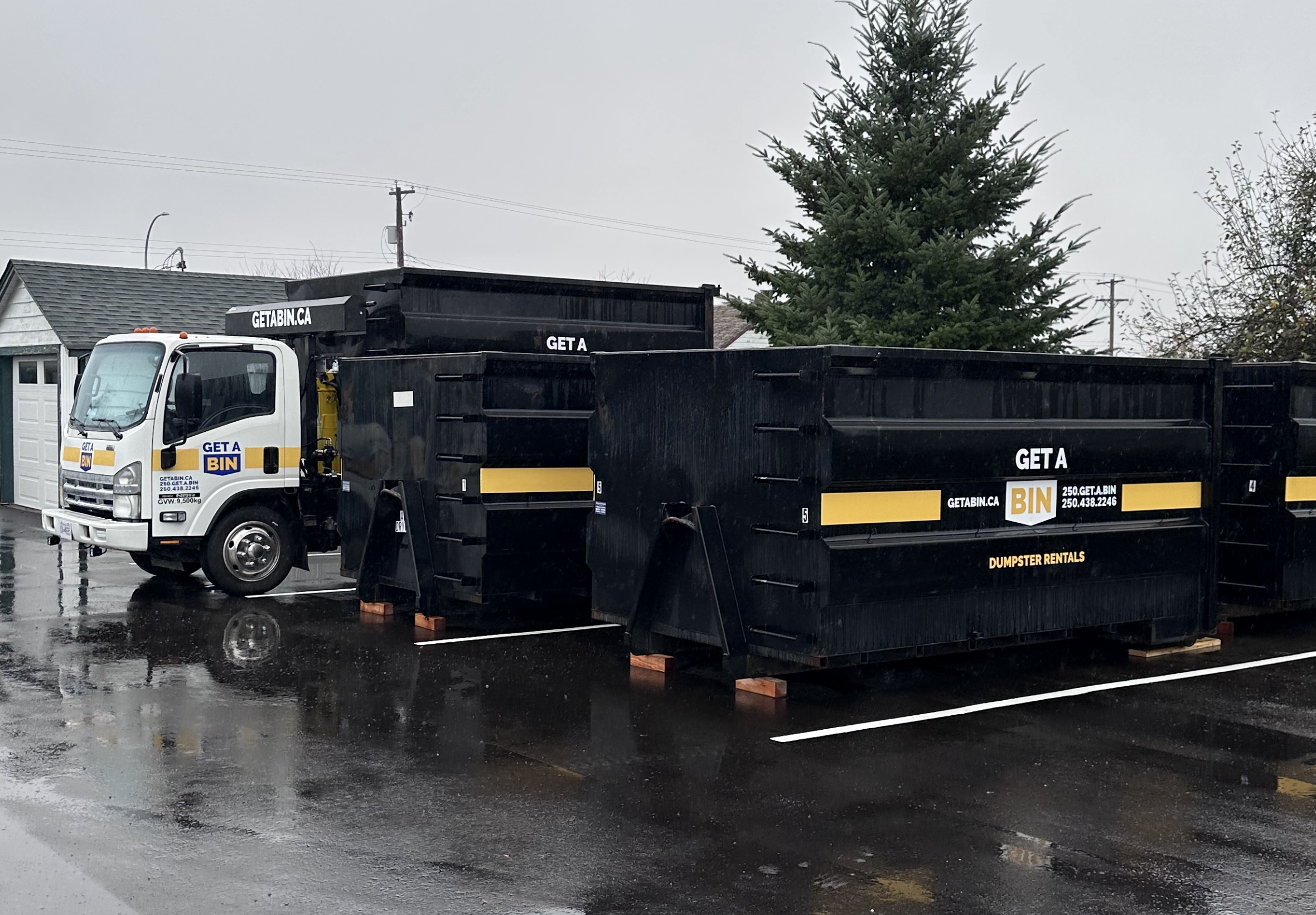 A white garbage truck with black and yellow storage containers parked on wet pavement in a residential area, with a large pine tree and utility poles in the background.