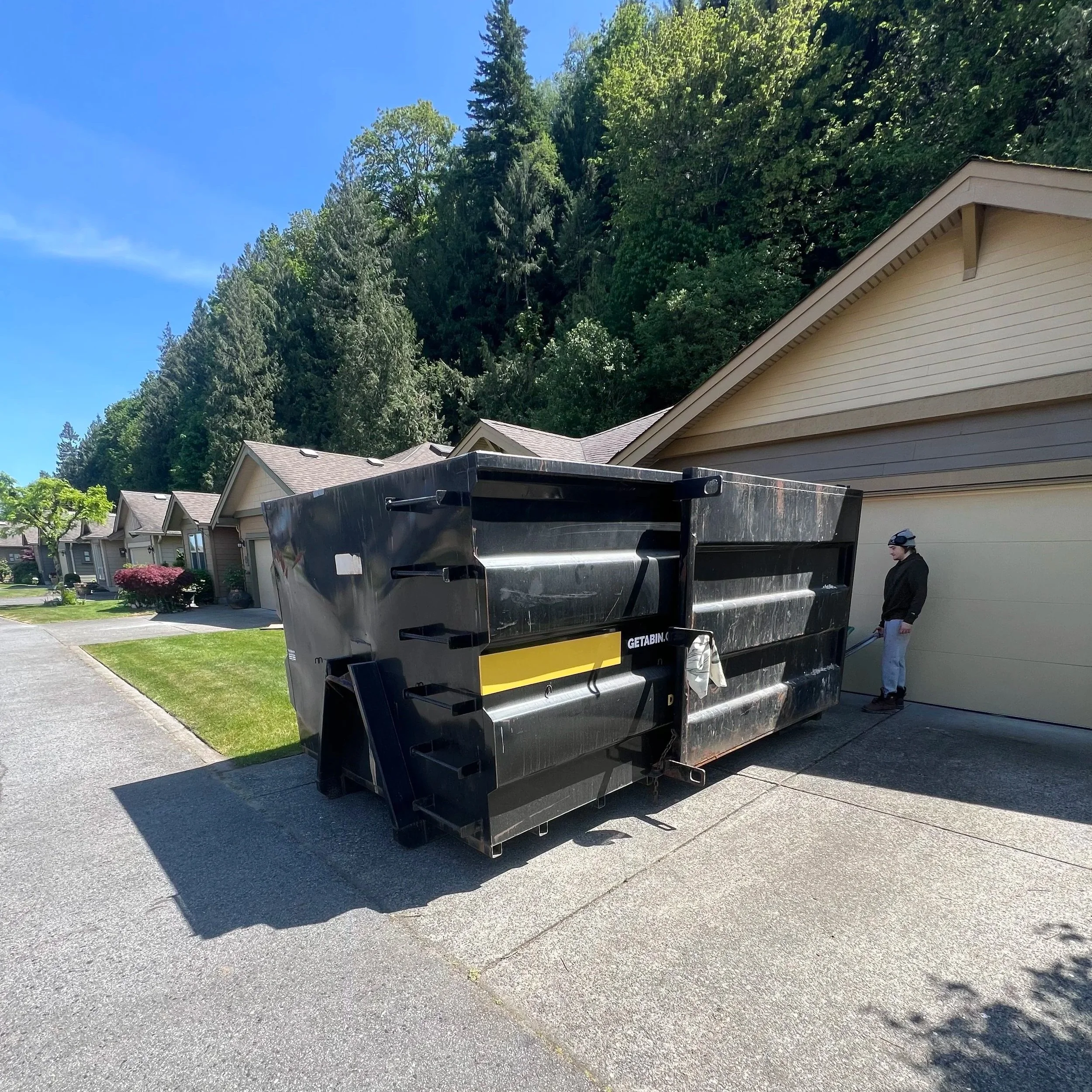 A residential neighborhood with row of houses, a large green hill covered with trees in the background, and a black trash dumpster in the driveway. There is a man standing near the dumpster. The sky is clear and blue.