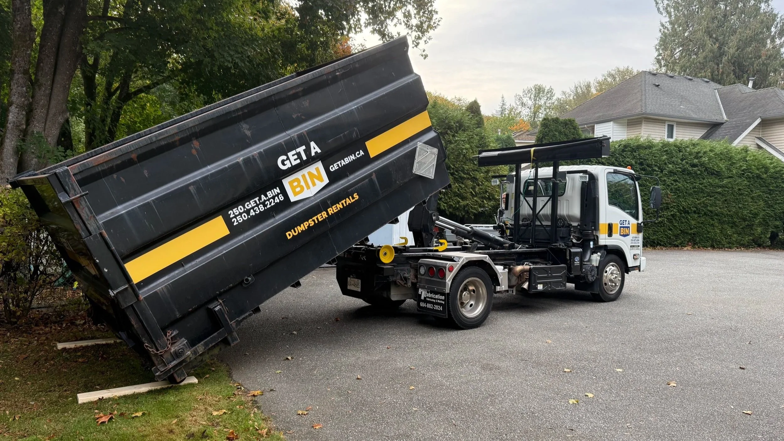 A dump truck with a black bed in a suburban neighborhood, lifting its bed to empty debris into the bed. The truck has a white cab with branding for GET A BIN and Dumpster Rentals, and is parked on the street near trees and houses.