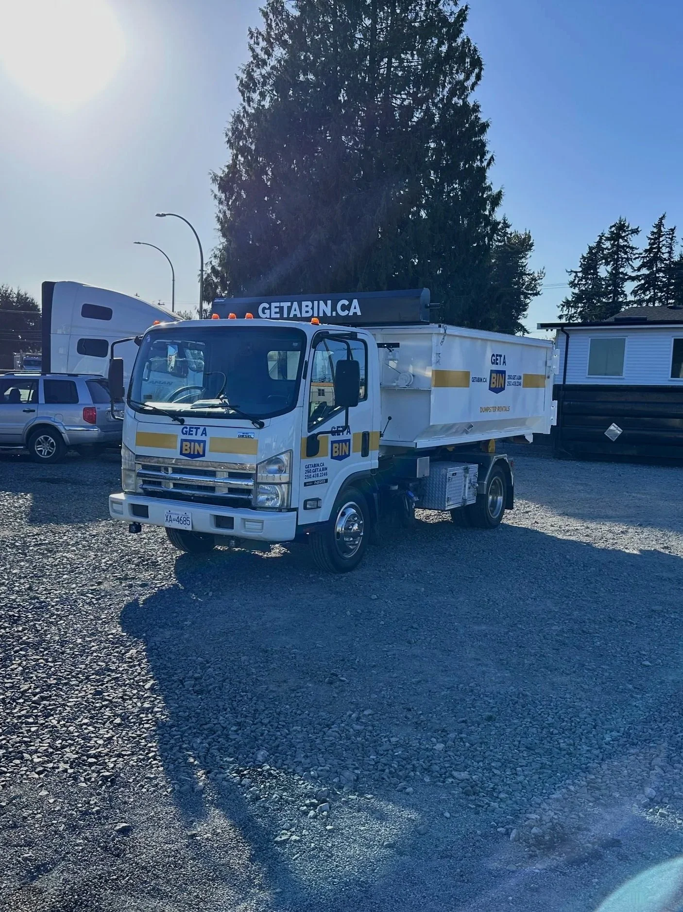 A white dumpster rental truck with orange warning lights on top, parked on a gravel lot, with the website GETABIN.CA displayed on a sign above the cab and the side of the container, which also has the text GETA BIN and contact information. There is a large tree and smaller trees in the background, along with a clear blue sky and a few other parked vehicles.