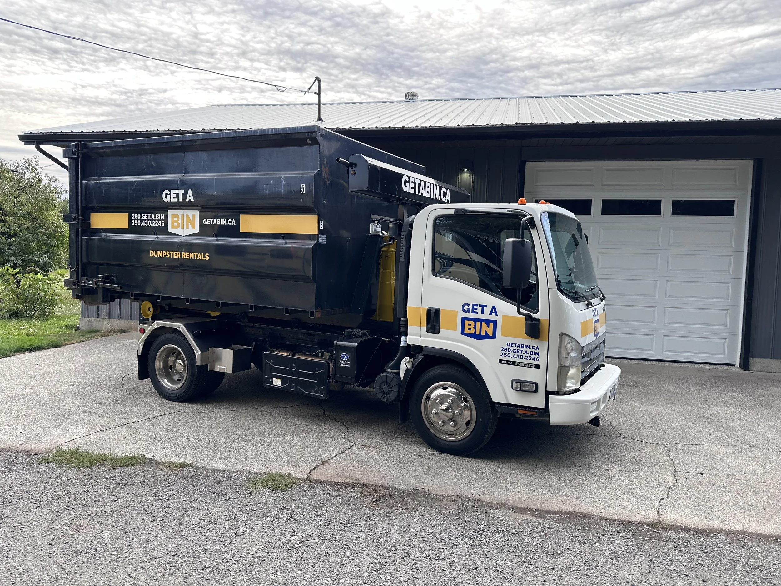 A white truck parked in front of a garage, with a black dumpster on the back that has white and yellow text reading 'GET A BIN' and court information, and a yellow stripe.