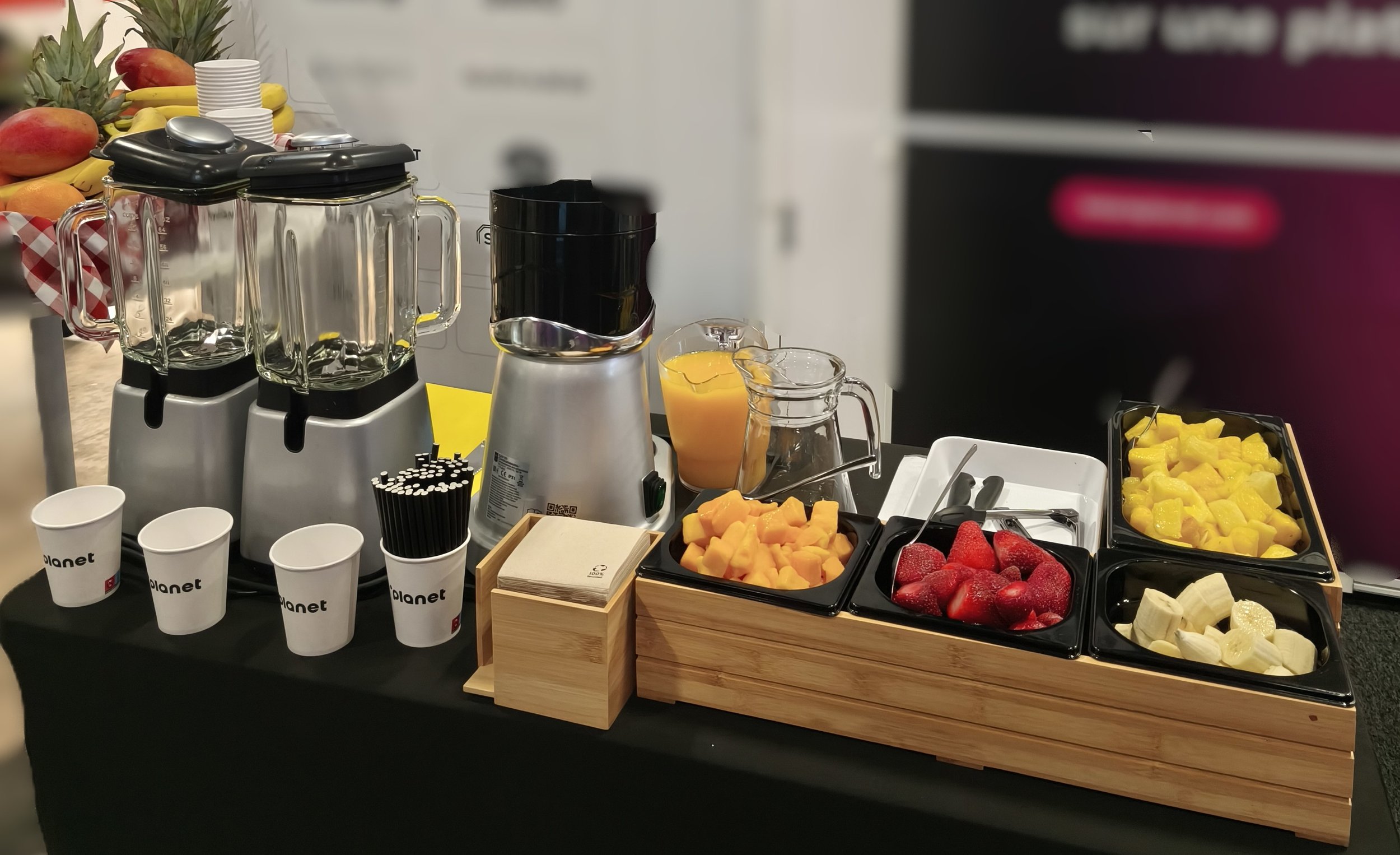 Station de boissons avec mixeurs, fruits frais, gobelets en papier, et conteneurs de fruits coupés comme fraises, mangues, bananes et ananas. smoothie, jus frais