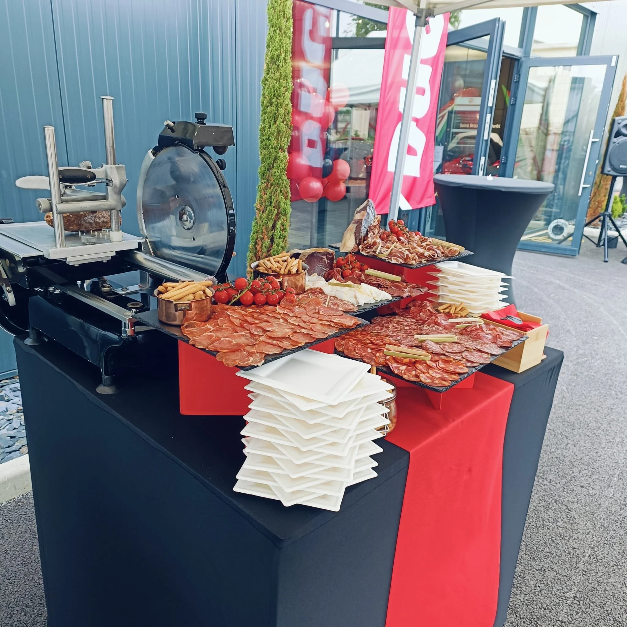 Buffet de charcuterie avec jambons, salamis, tomates cerises, et crackers, devant une trancheuse à viande, à l'extérieur. pata negra