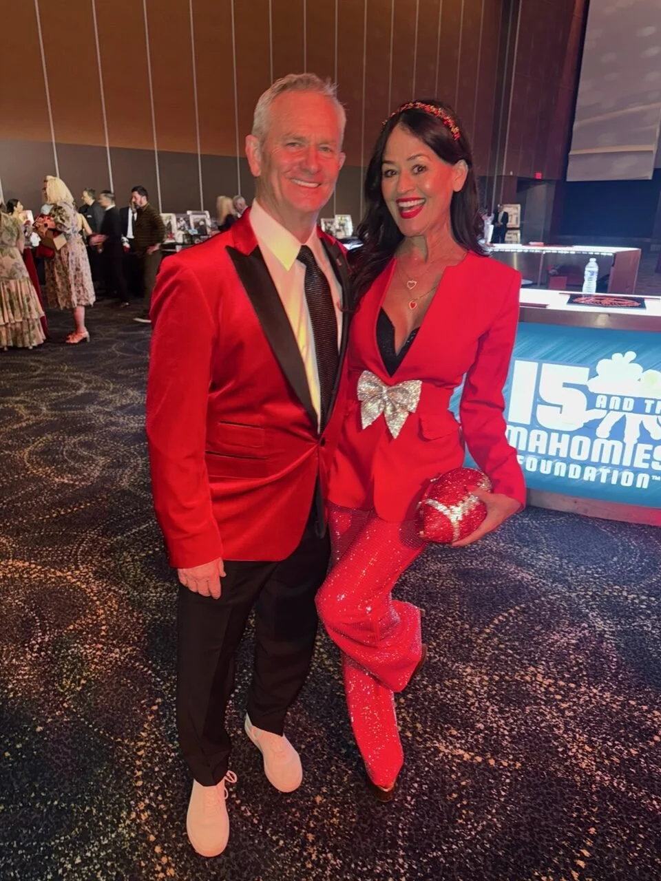 A man and Yve Rojas dressed in red at a formal event. The woman is holding a sparkling red clutch and is wearing a matching red outfit with sequined pants and a heart necklace.